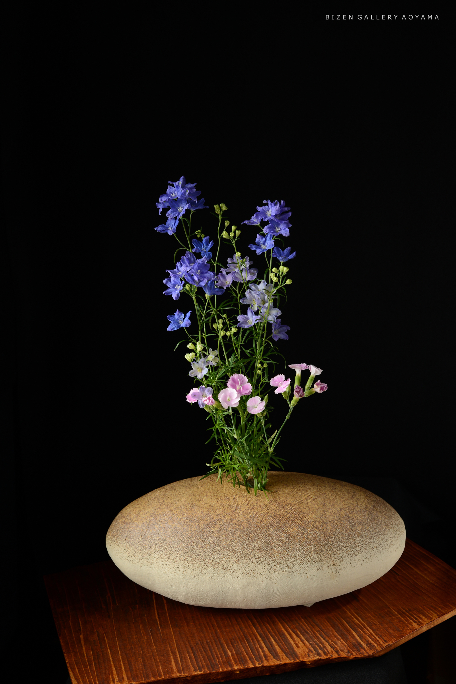 黒い背景に美しい青紫色の花と薄いピンクの花が生けられた陶器の花器の画像。