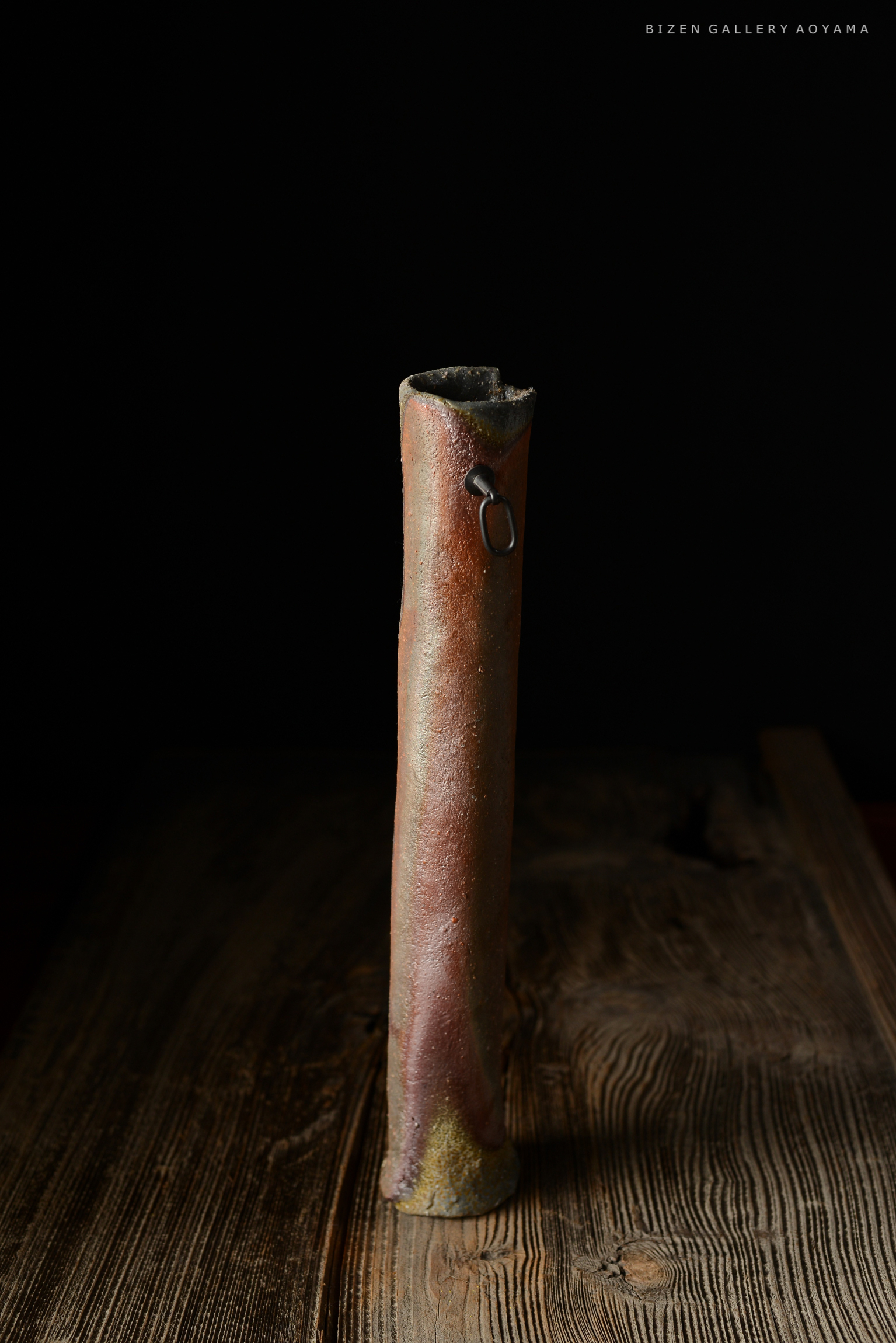 暗い背景の前に立てられた陶器の花瓶。円筒形で、側面には金属のリングが付いている。