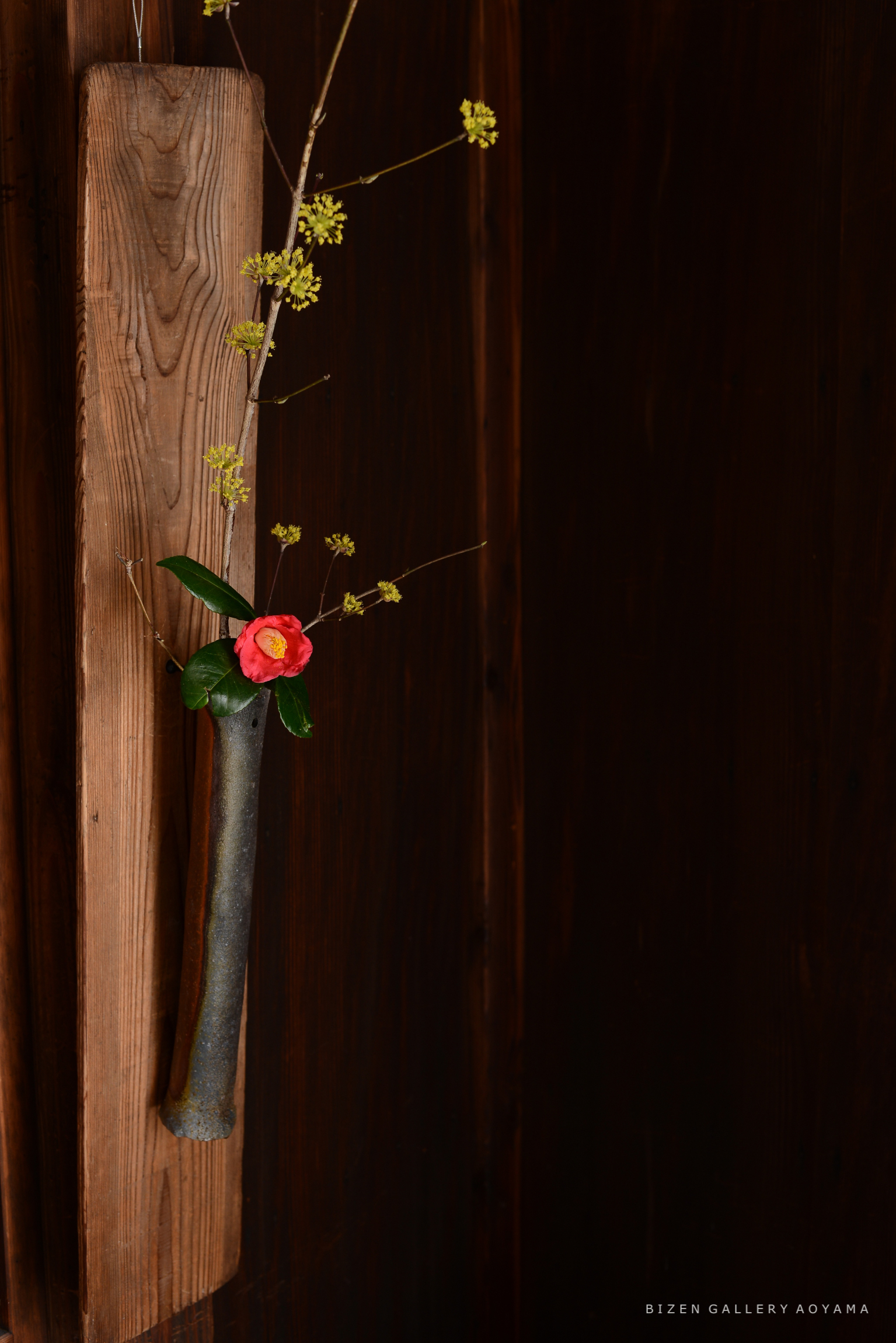 黒い陶器の花瓶に花が生けられている様子。背景は木の壁で、一部には木の板が見える。