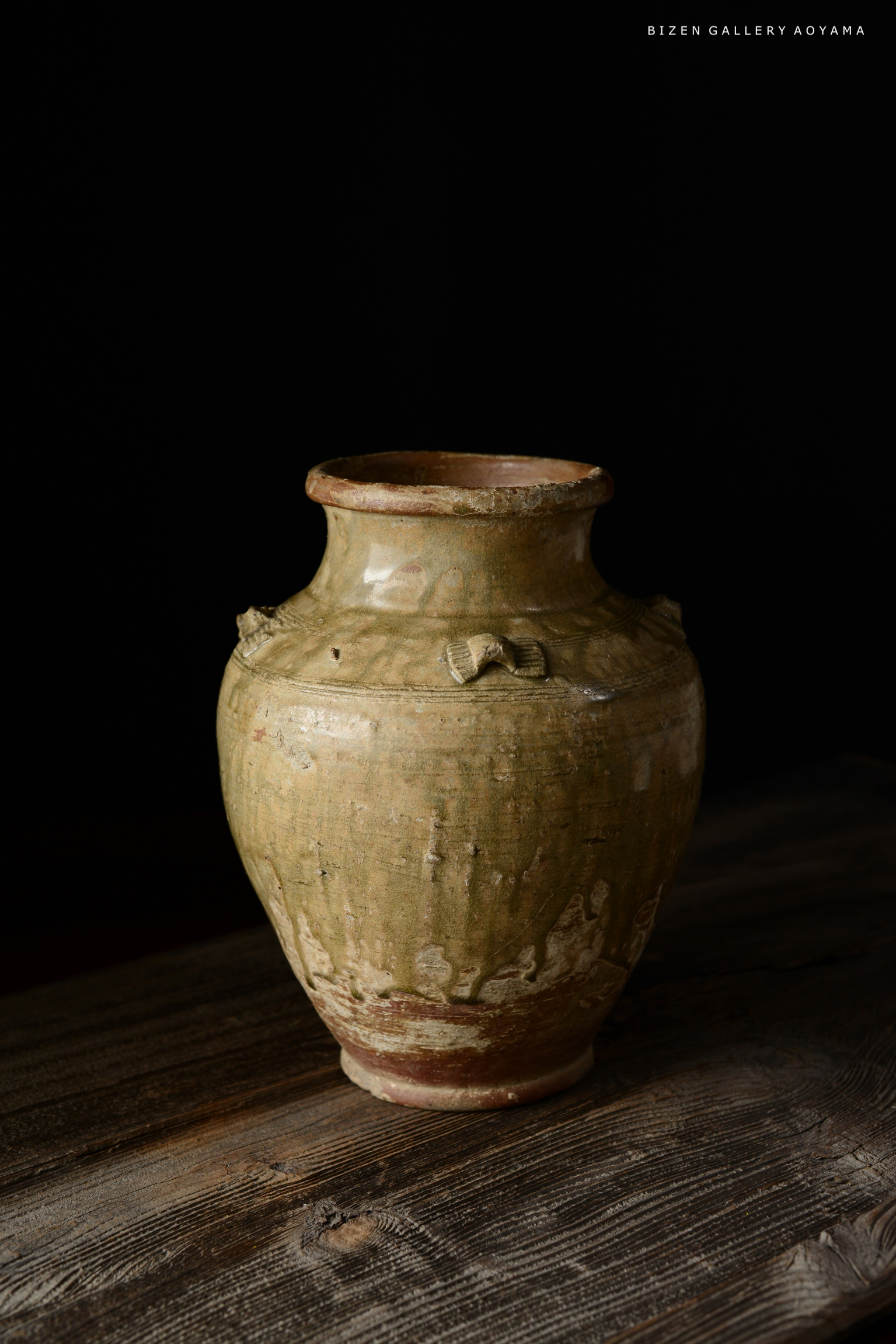 緑がかった陶器の壺が木製のテーブルの上に置かれている。背景は黒で、壺の形状やテクスチャーが際立っている。