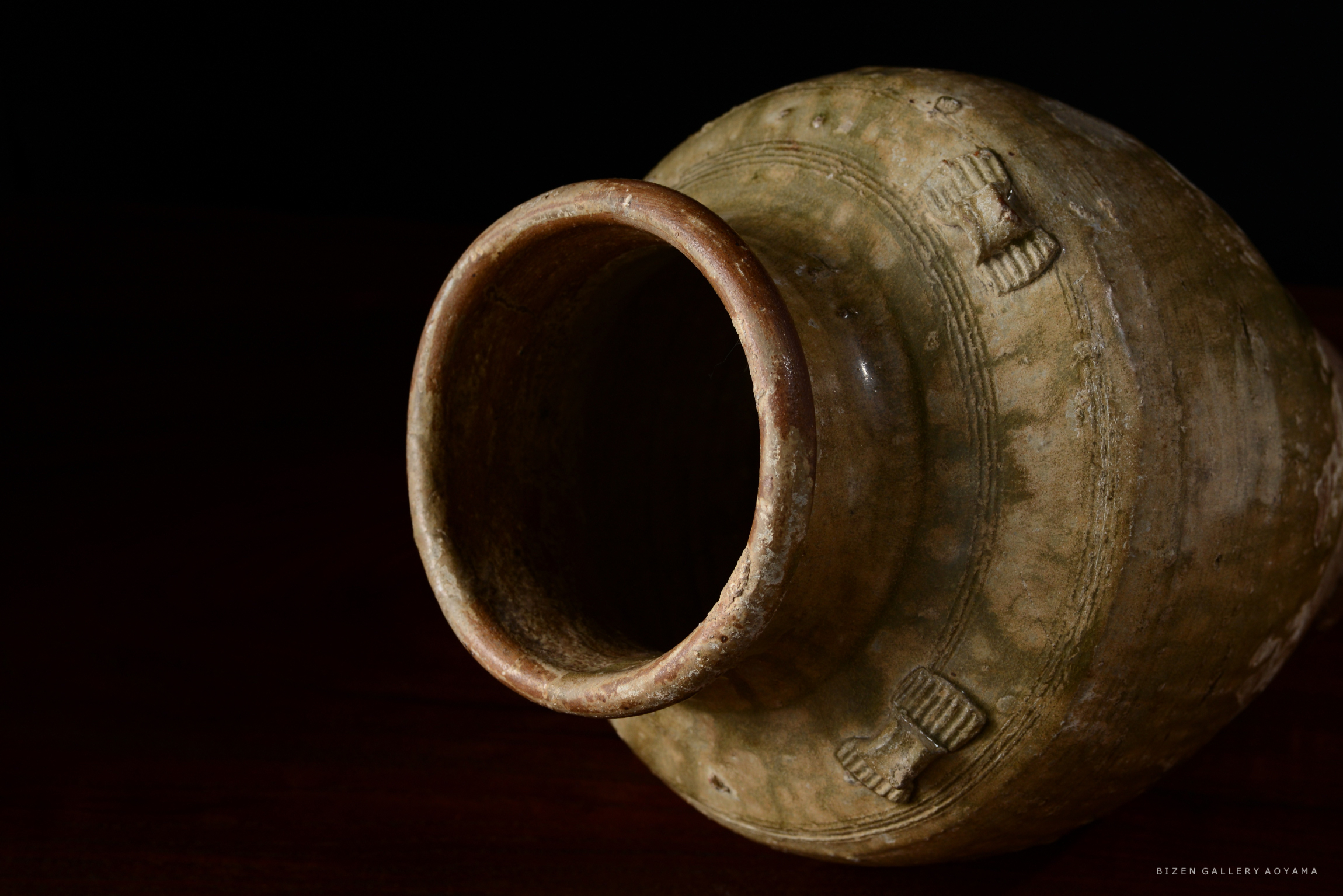 陶器の壺が横向きに置かれている。壺の口が手前にあり、茶色の地味な模様が施されている。