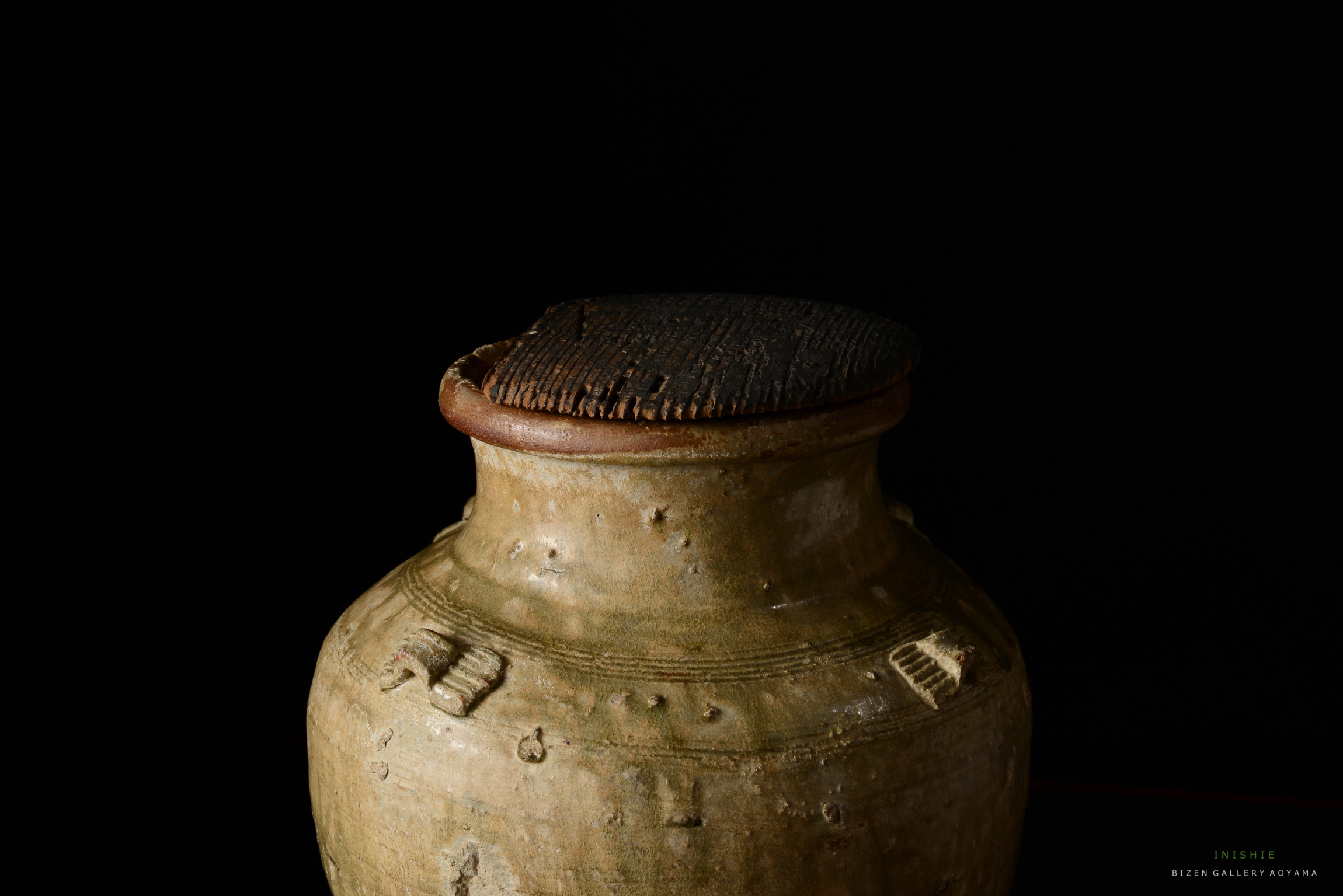 黒い背景の前に置かれた、陶器の壺の上部の画像。壺は自然な色合いで、粗い表面と特徴的な蓋を持っています。