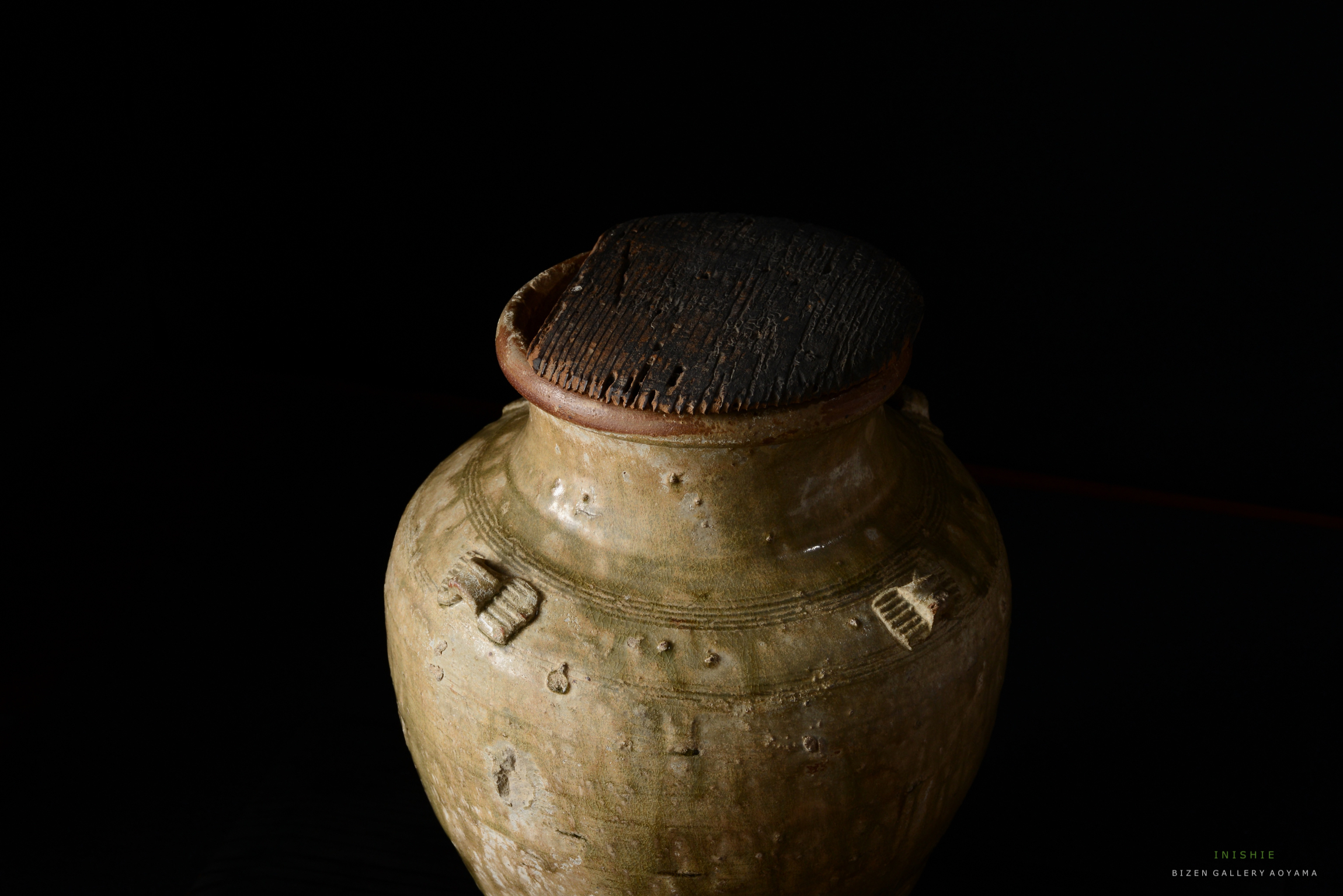 暗い背景に置かれた、土製の壺の画像。蓋は木製で、表面には線が見える。壺の側面には装飾的な模様が施されている。
