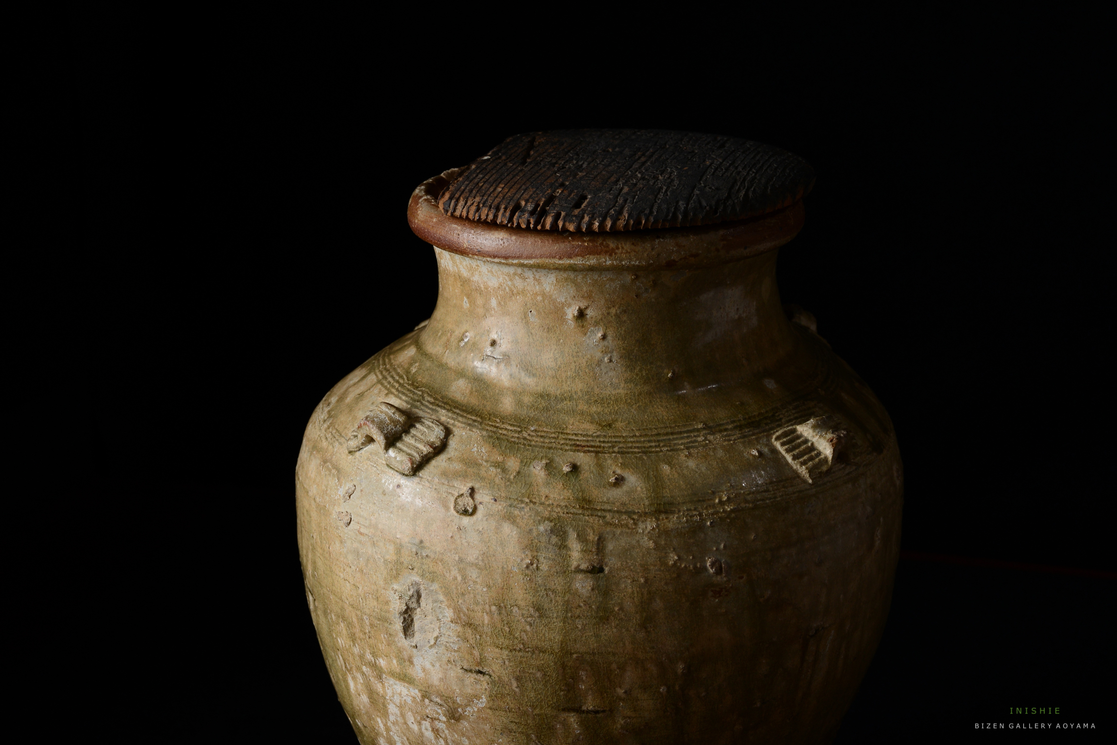 黒背景に置かれた、伝統的な日本の陶器の壺。壺の上部には木製のふたがあり、表面には彫刻の模様が見える。