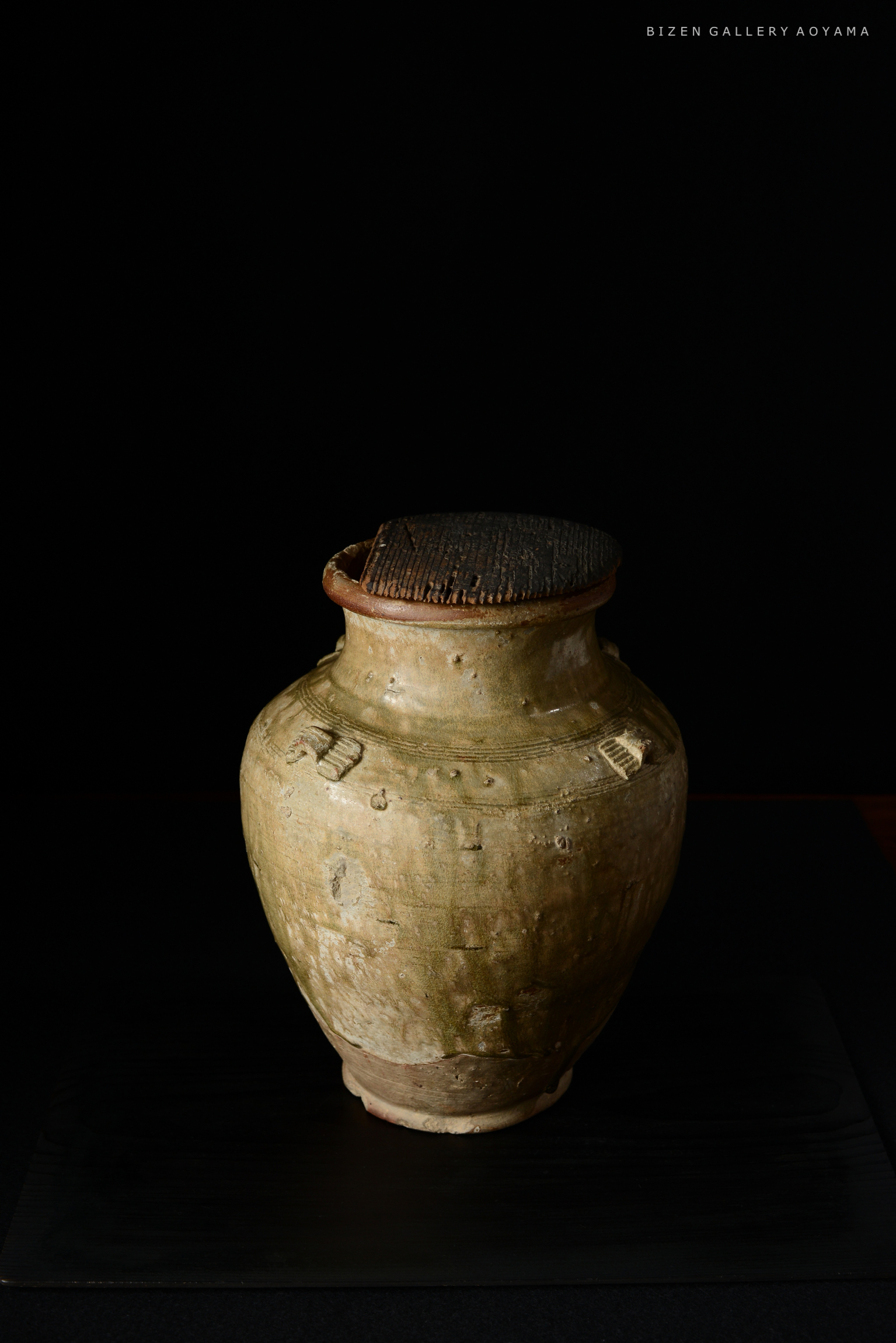 黒い背景に置かれた古い陶器の壷。壷の上には板状の蓋があり、表面には細かい彫刻が施されている。