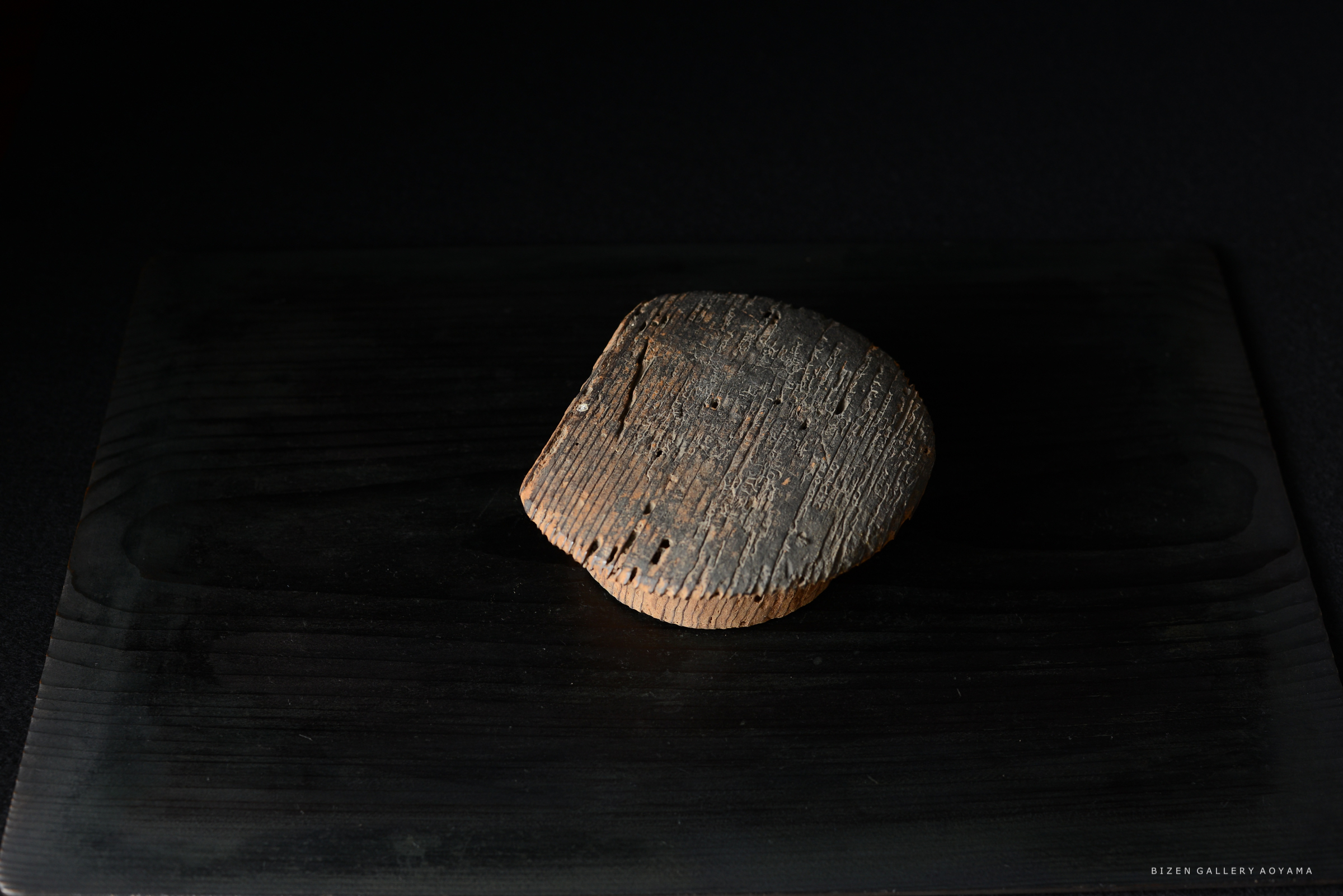 黒い背景の上に置かれた、古い土器の一部が映っている。土器は剥がれた部分や模様が見受けられ、独特の形状をしている。