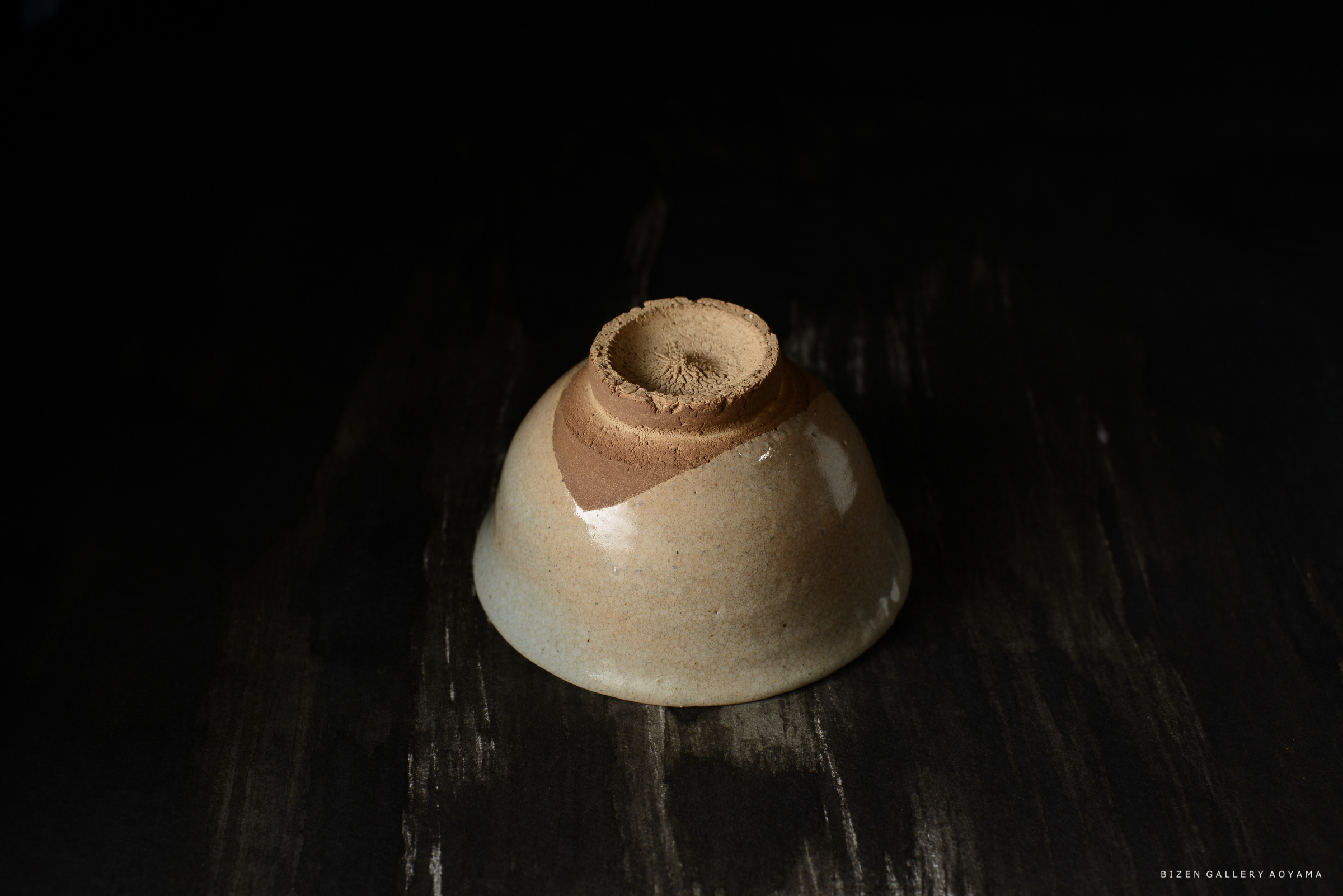 黒い背景の上に置かれた、ユニークなデザインの陶器の器。上部は滑らかで艶やかな表面で、色はクリーム色と茶色のグラデーション。