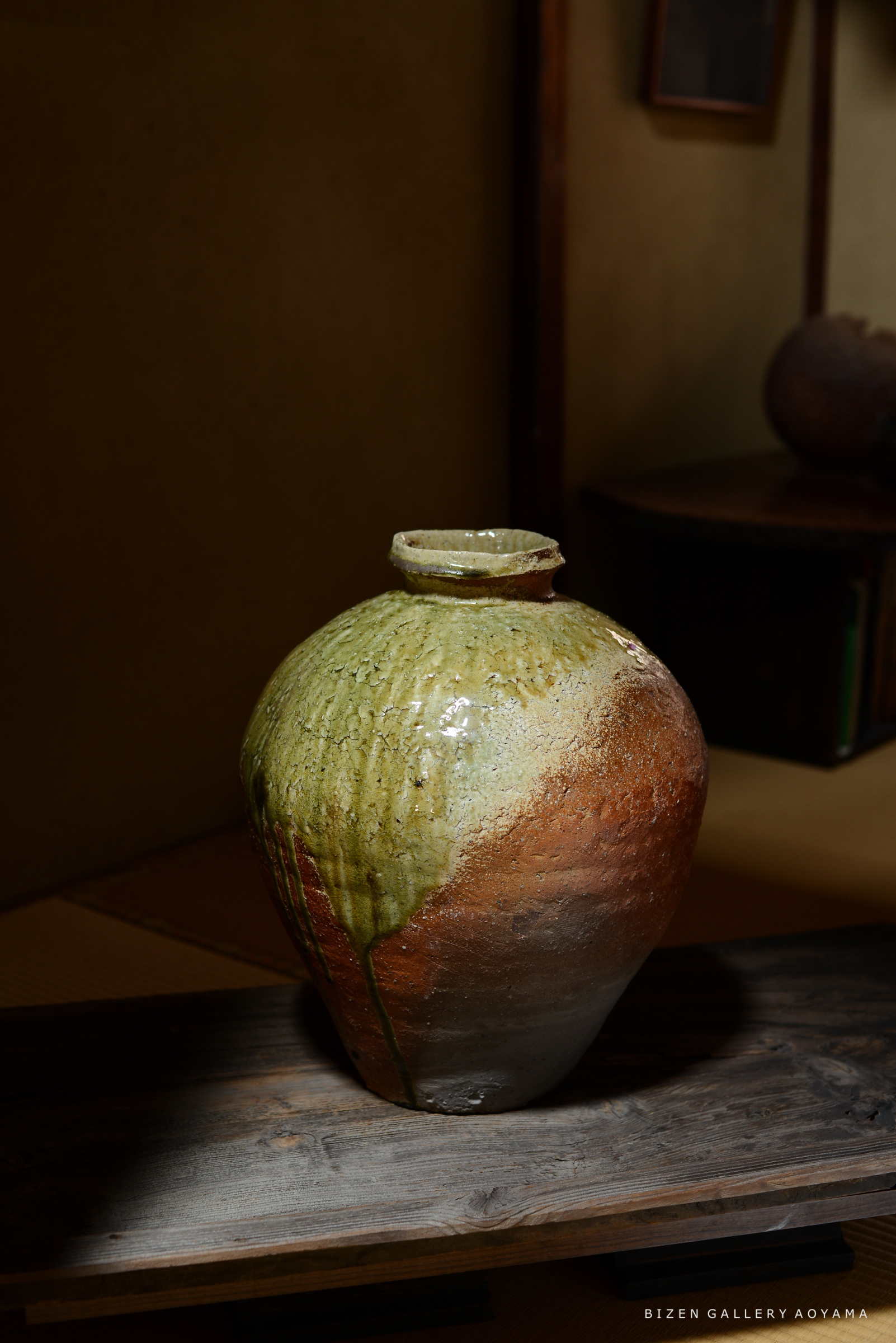 陶器の壺が木の台の上に置かれている。底部は灰色で、上部は緑と茶色のグラデーションが特徴的。背景は和室の雰囲気が感じられる。