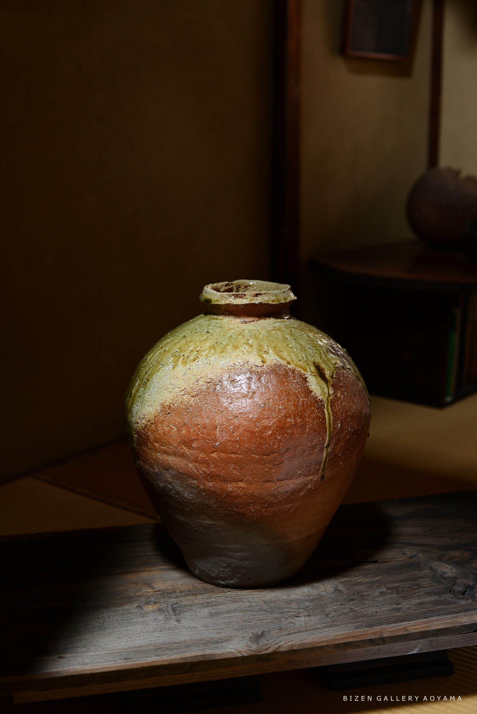 美しい釉薬が施された大きな陶器の壺が、木製の台の上に置かれている。背景には和室の内装が見える。