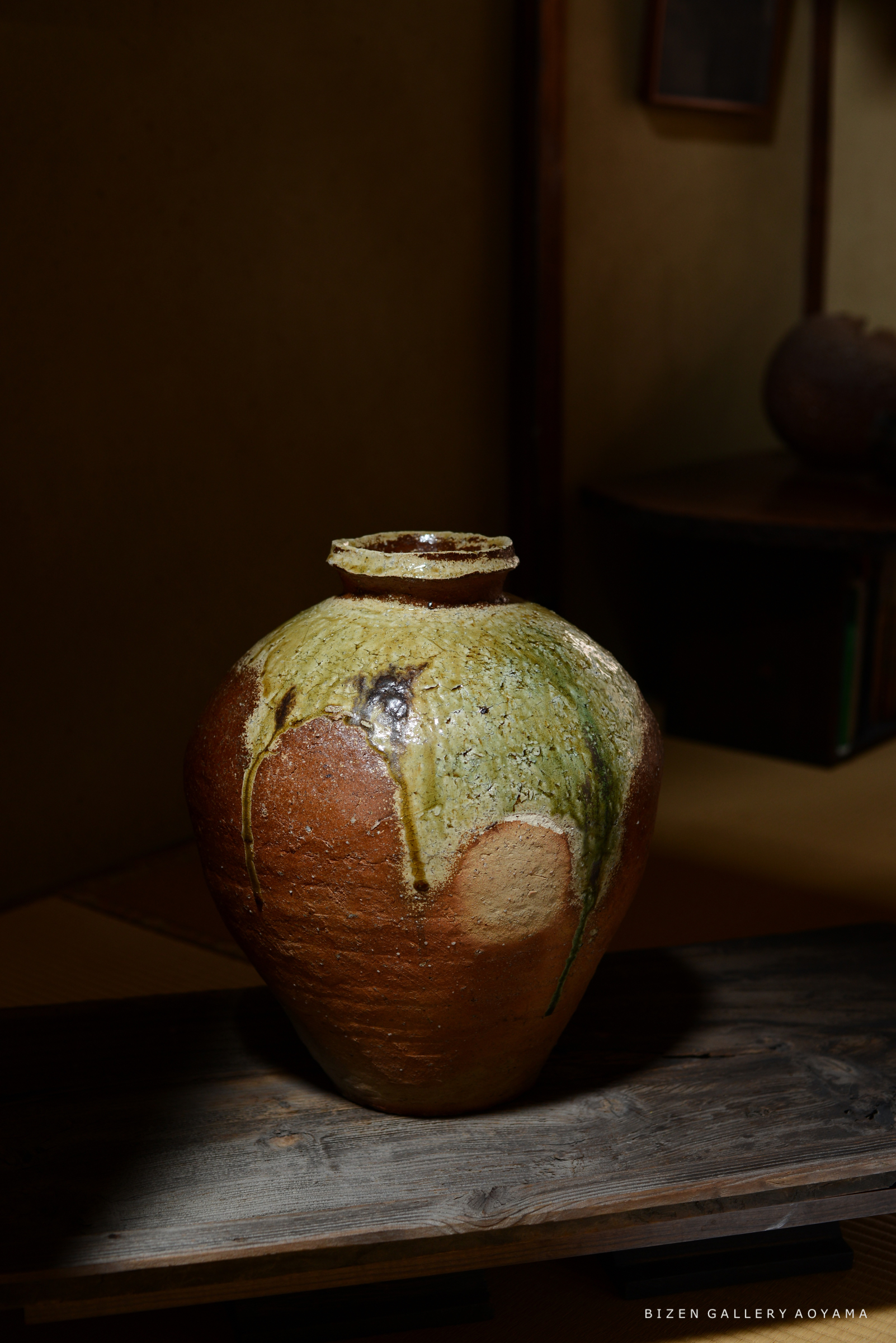 光の中に置かれた美しい陶器の壺。上部は緑がかった釉薬で装飾され、下部は温かみのある茶色。木の台の上に置かれている。