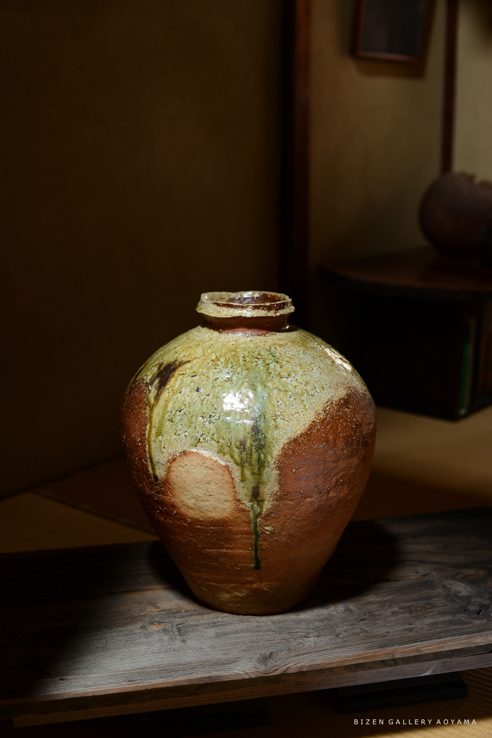 ビゼン焼の陶器が木の台の上に置かれている画像。陶器は茶色と緑色の釉薬が施されており、独特の形状を持っている。背景には壁と家具の一部がぼんやりと見える。