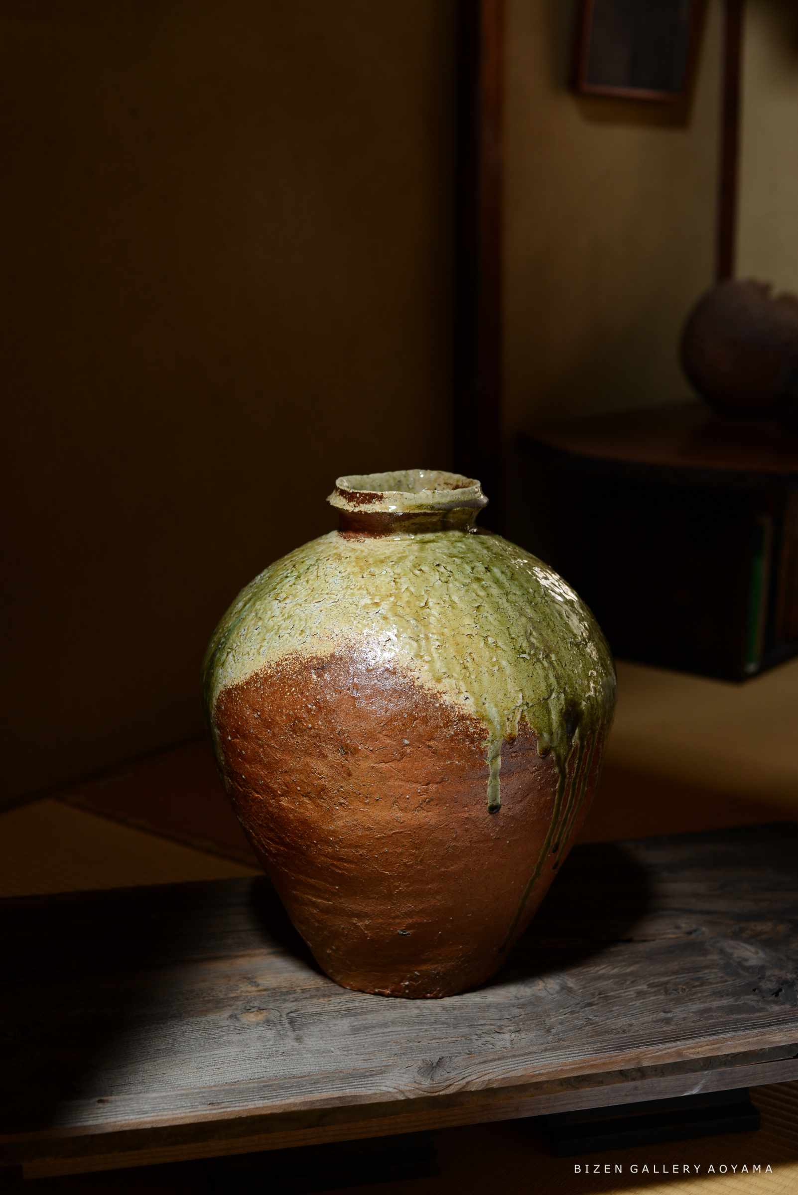 緑色と赤茶色の釉薬が施された陶器の瓶が、木製の台の上に置かれている。背景はシンプルで、温かみのある雰囲気を醸し出している。