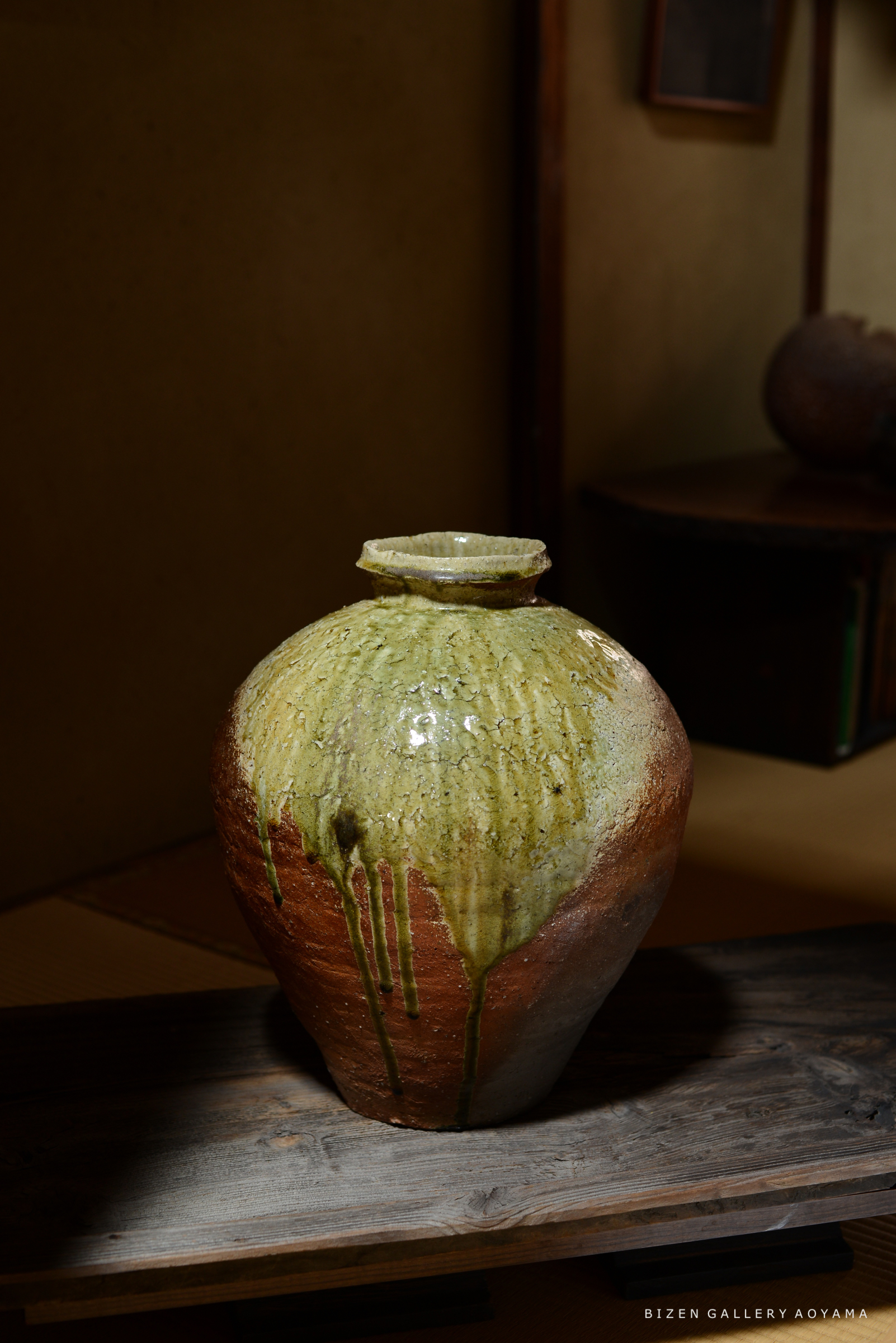 緑色の釉薬がかかった陶器の壺が、テーブルの上に置かれています。壺の下部は茶色で、釉薬が滴り落ちた跡が見えます。背景には和室の壁と他の陶器がわずかに映っています。