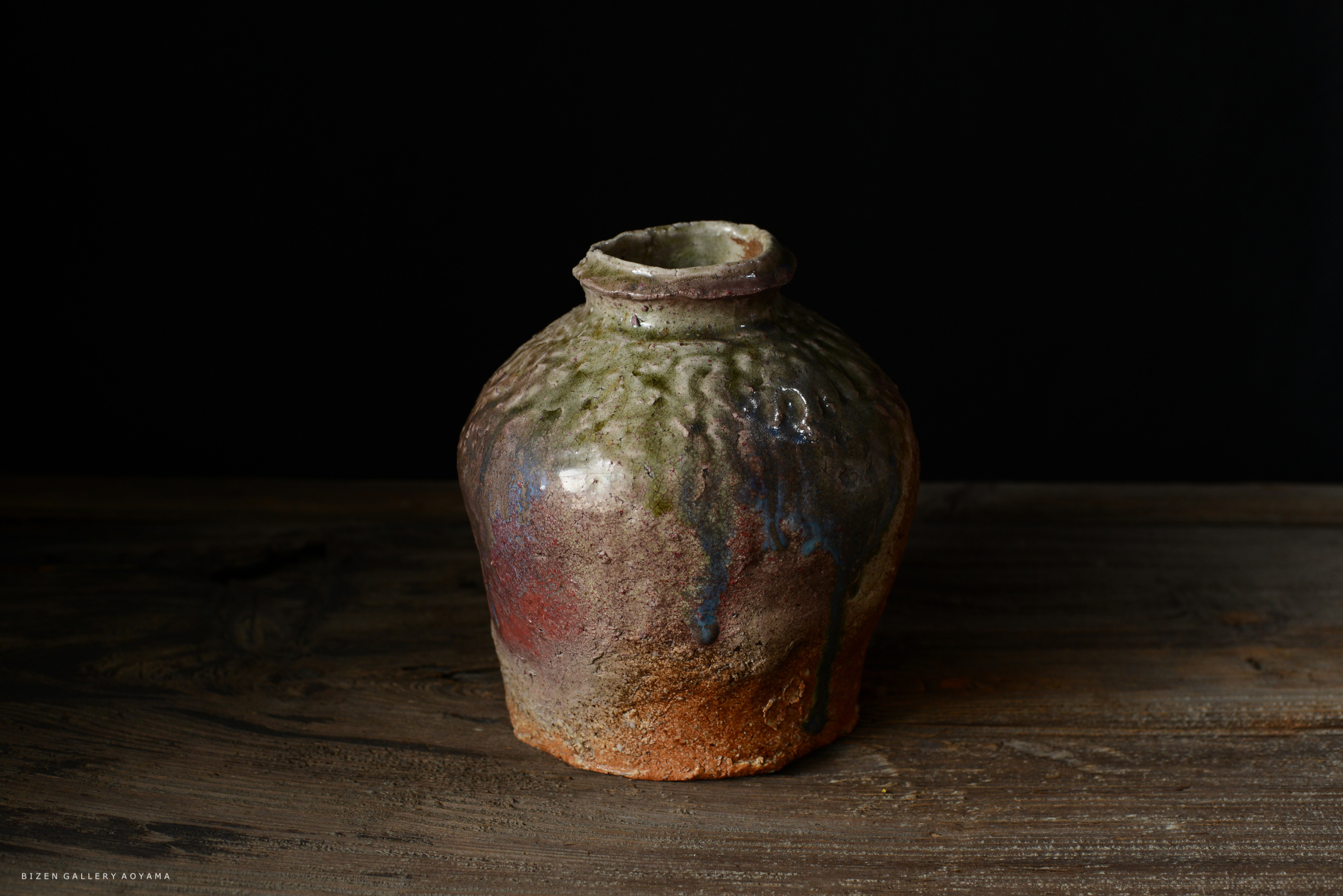 色とりどりの釉薬が施された陶器の壺。木製のテーブルの上に置かれ、暗い背景が特色を際立たせている。