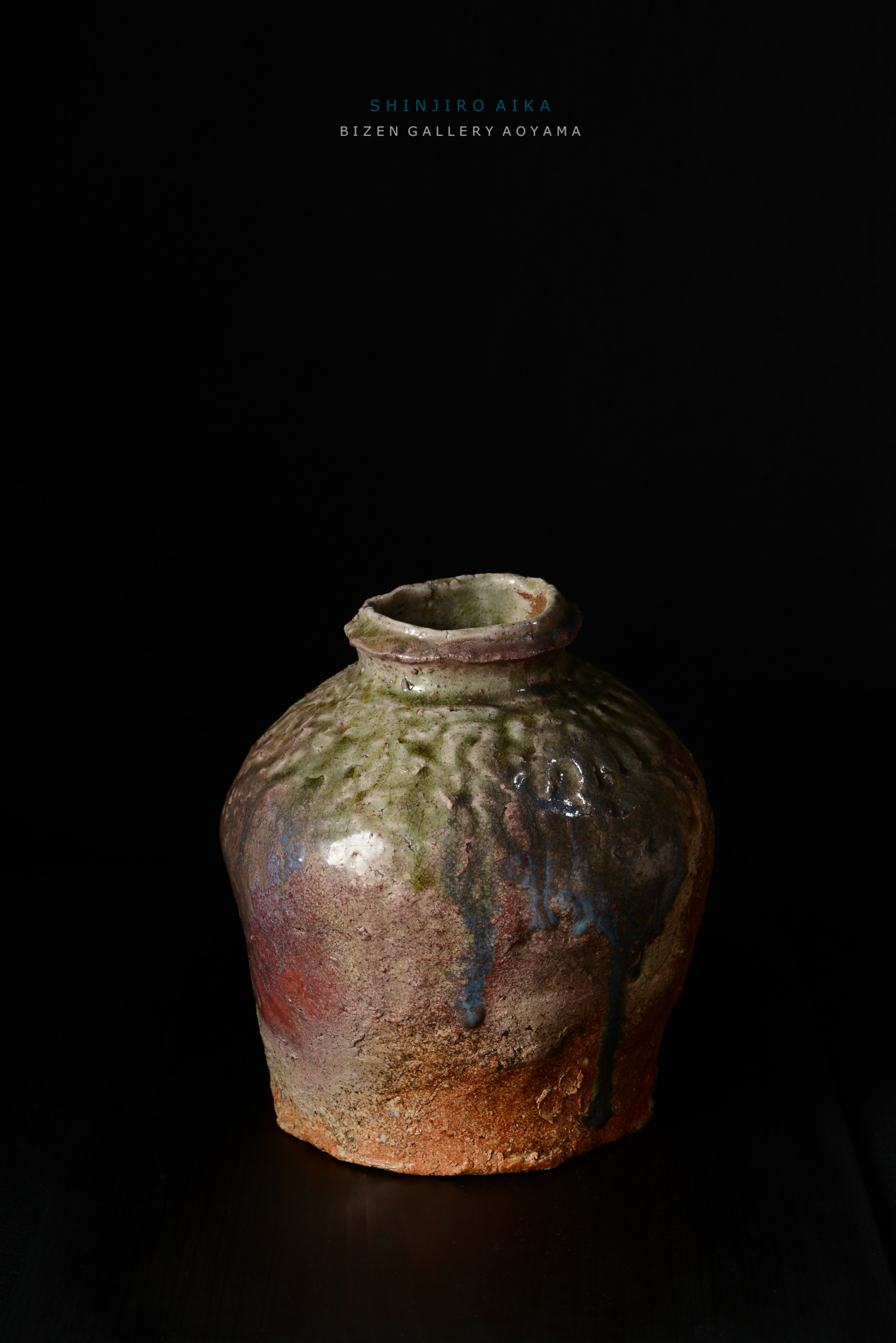 黒い背景の前に置かれた、独特の形状を持つ陶器の花瓶。表面には色褪せた緑や赤の釉薬が施され、ところどころに滴り落ちたような模様が見られる。