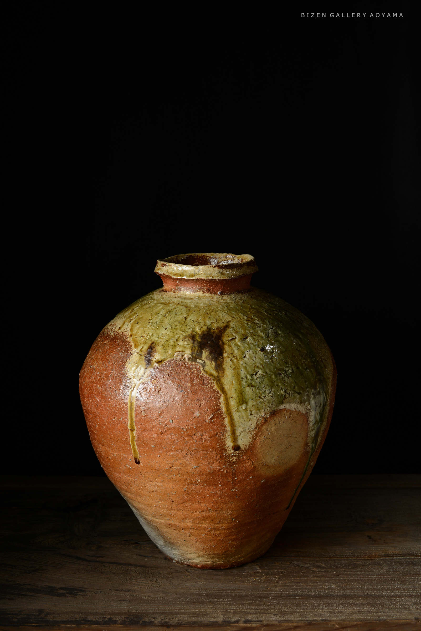 黒背景の前に置かれた、陶製の壺。茶色と緑の釉薬が施されており、独特の模様やテクスチャが見られる。
