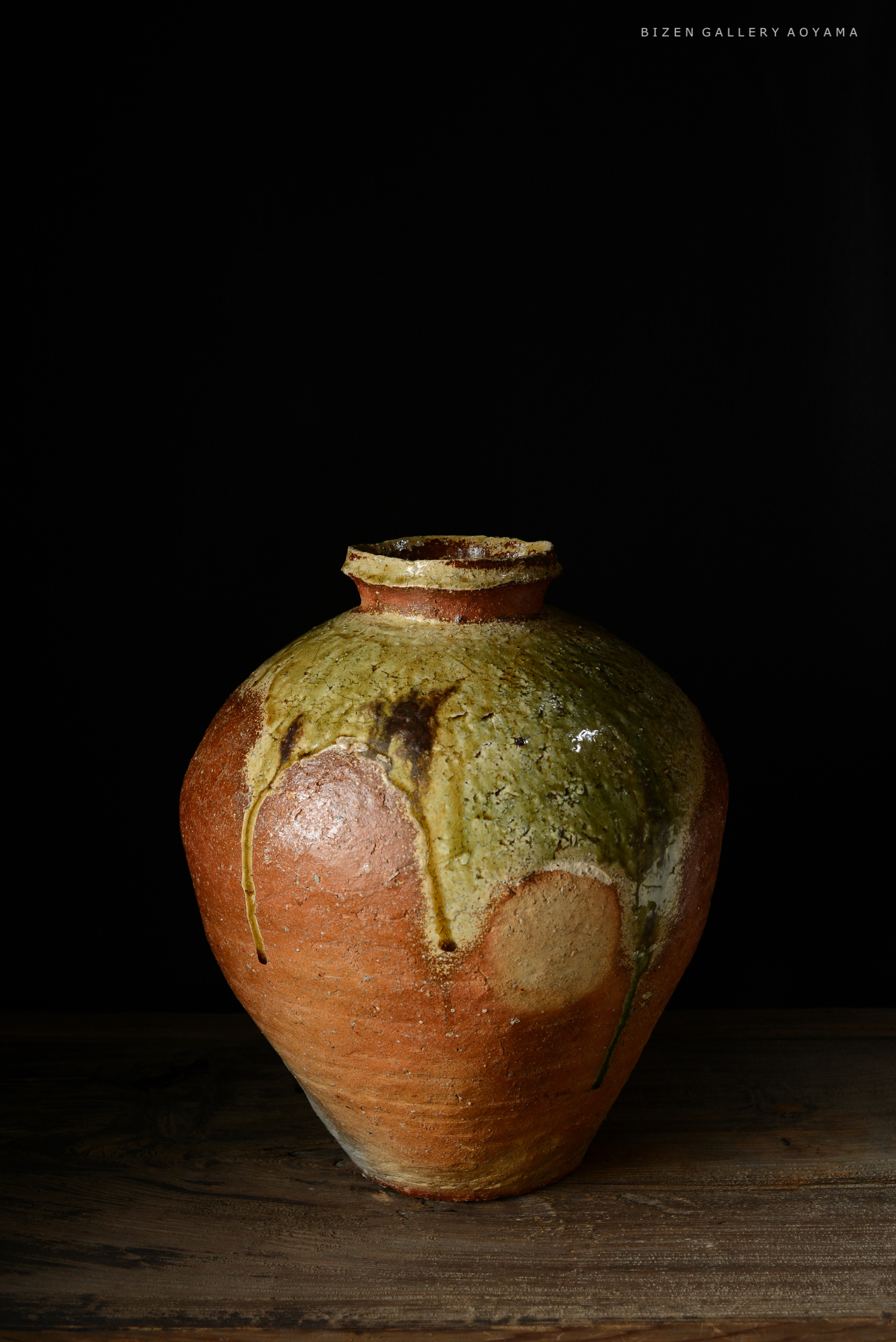 黒い背景の前に置かれた、緑と茶色の釉薬が施されたビゼン焼きの陶器の壺