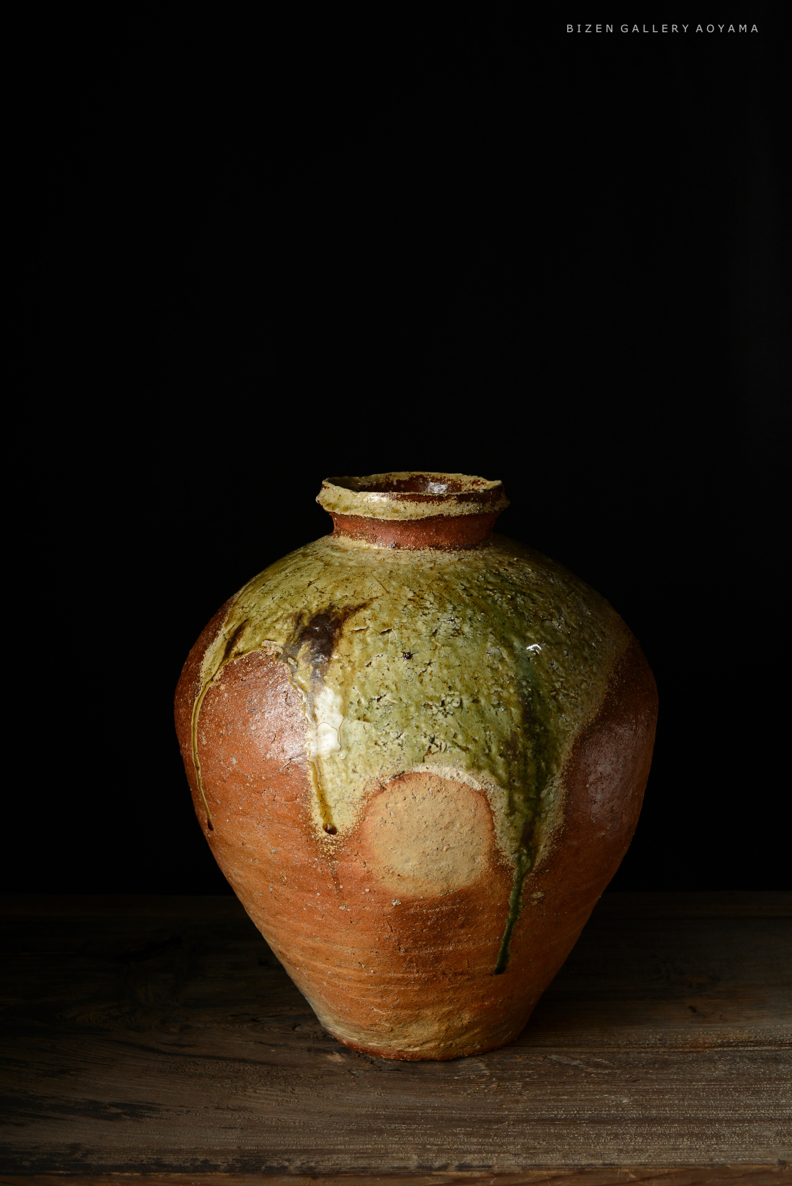 黒い背景を背にした、土でできた独特の形状の壺。上部には緑色の釉薬がかかっており、自然な風合いが感じられる。