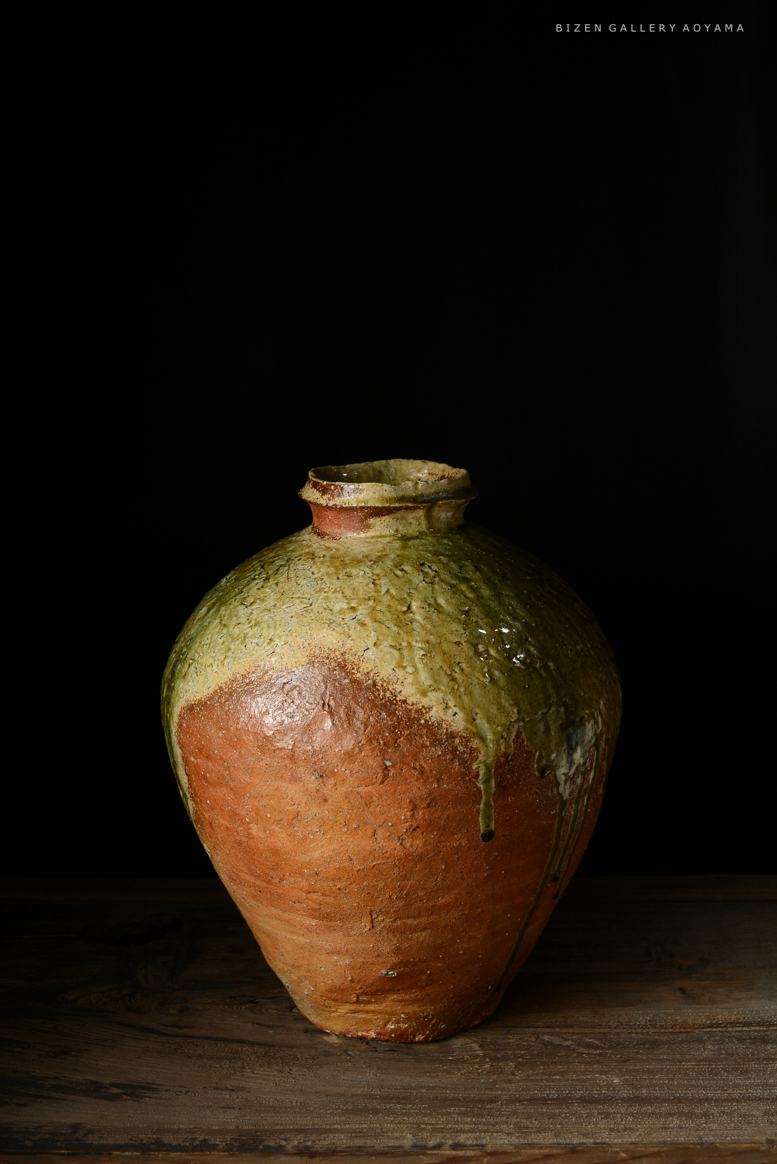 黒い背景の前に置かれた、上部が緑で下部がオレンジのビゼン焼きの壺