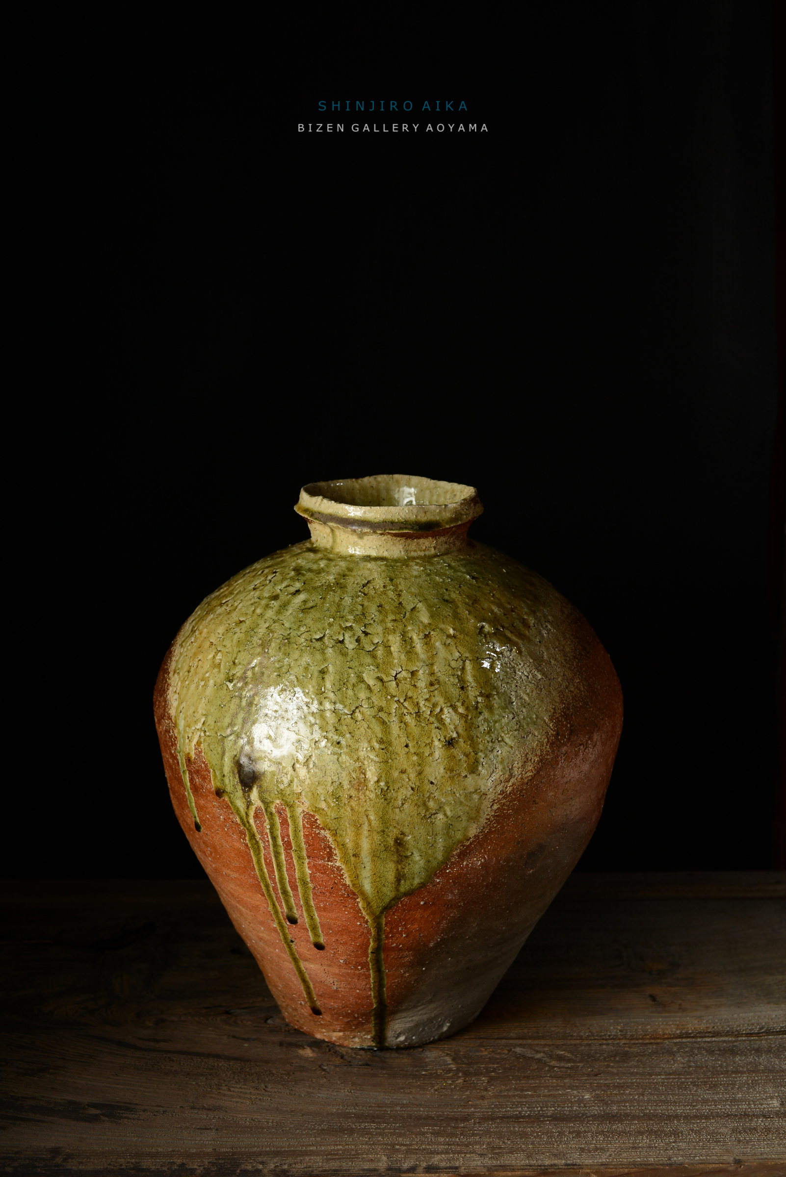 緑色の釉薬がかかった陶器の壺が木のテーブルの上に置かれています。壺は、下部がオレンジ色で、質感が豊かです。黒い背景が強調されています。