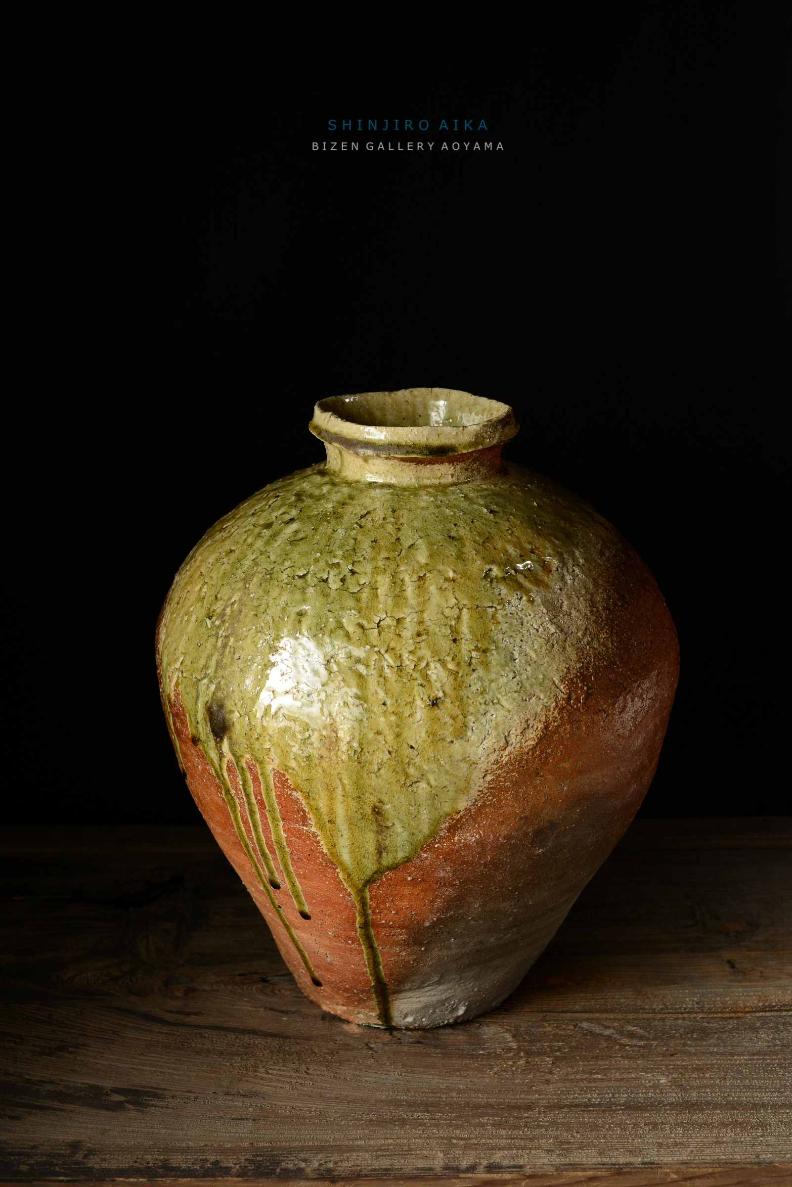 緑とオレンジの釉薬が施されたビゼン焼きの花瓶。滑らかな表面と独特な色合いが特徴。暗い背景の上に置かれた木製のテーブルの上にある。