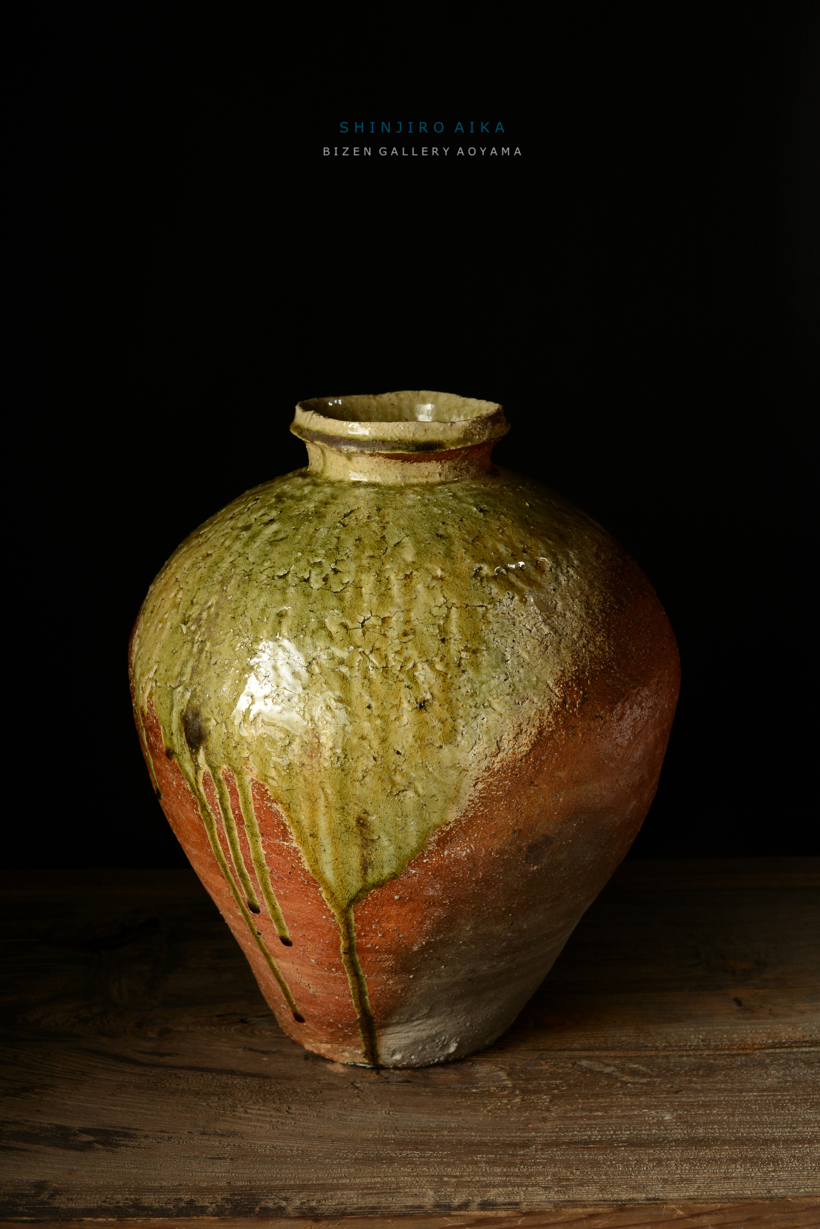 緑と茶色の釉薬が流れ落ちたビゼン焼きの壺。木のテーブルの上に置かれ、黒い背景が印象を引き立てている。