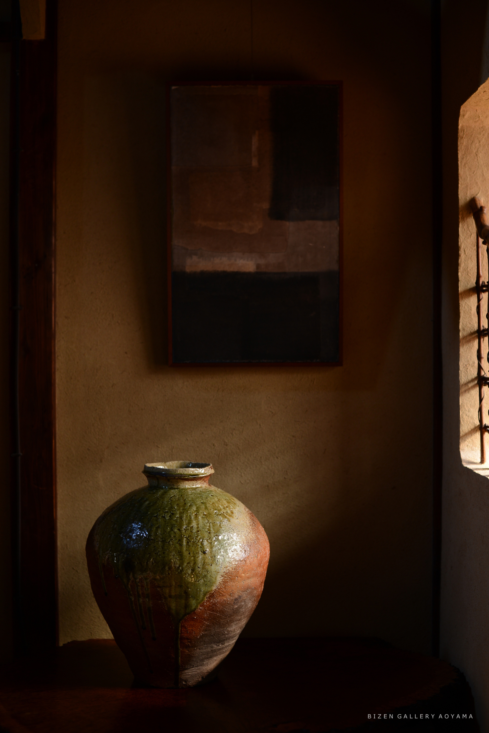 薄暗い部屋の中に置かれた、緑色の釉薬がかかったビゼン焼きの大きな花瓶と、その背後に掛けられた抽象画。