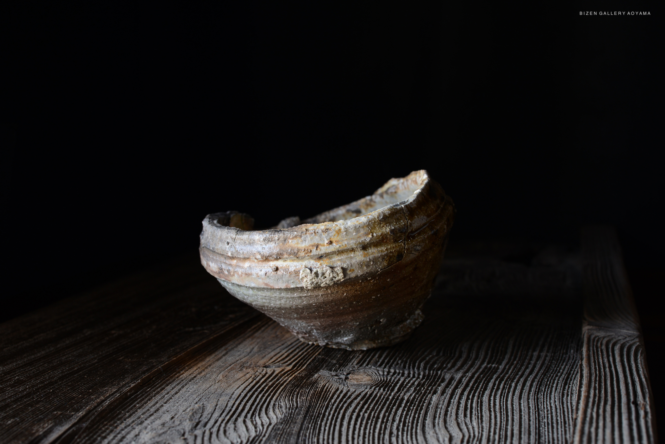 木のテーブルの上に置かれた、古い焼き物の器。暗い背景に対して、特徴的な形状とテクスチャが際立っている。