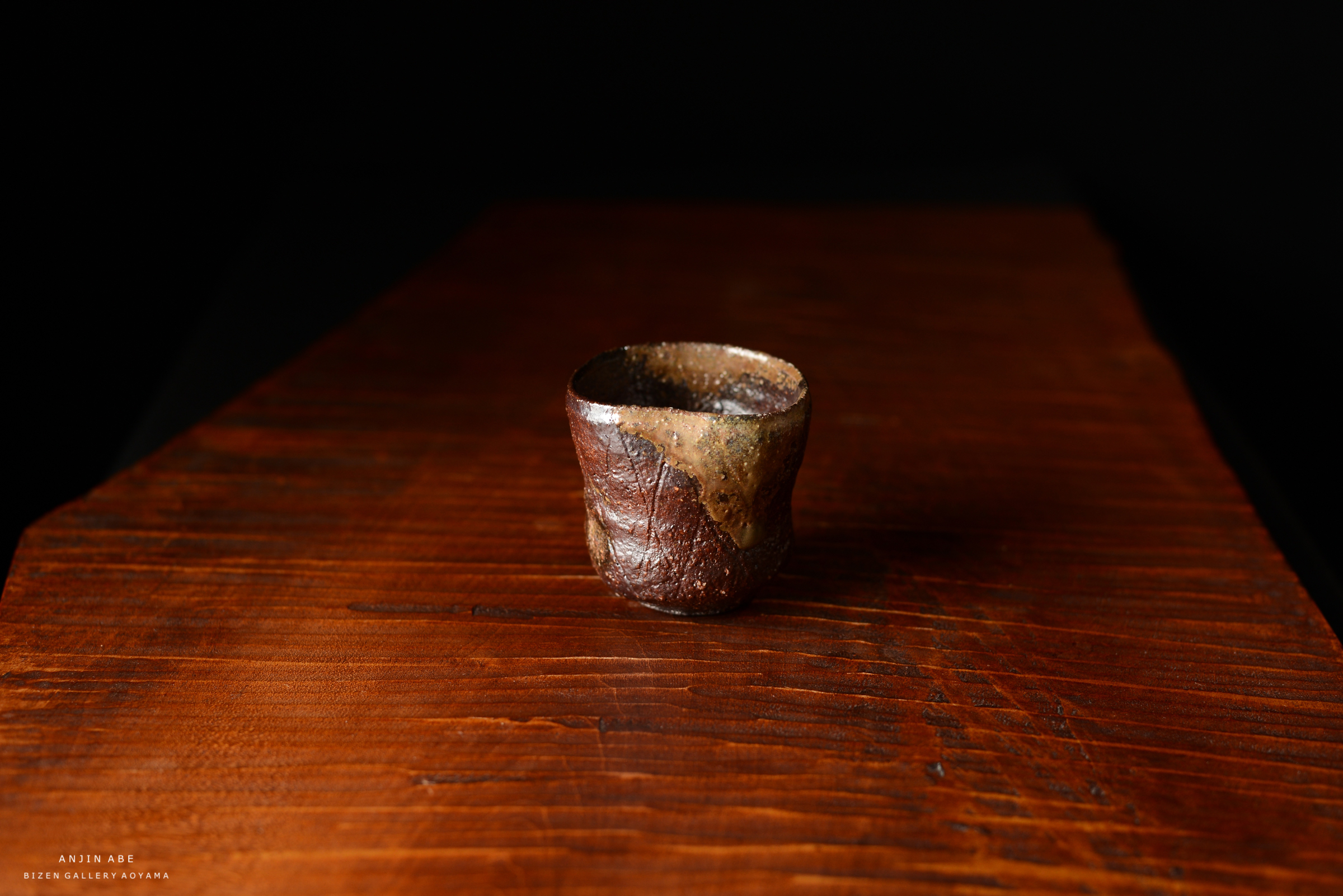 黒い背景の上に置かれた独特の形状の陶器のカップ。木のテーブルの上にあり、カップは土色の釉薬がかかっている。