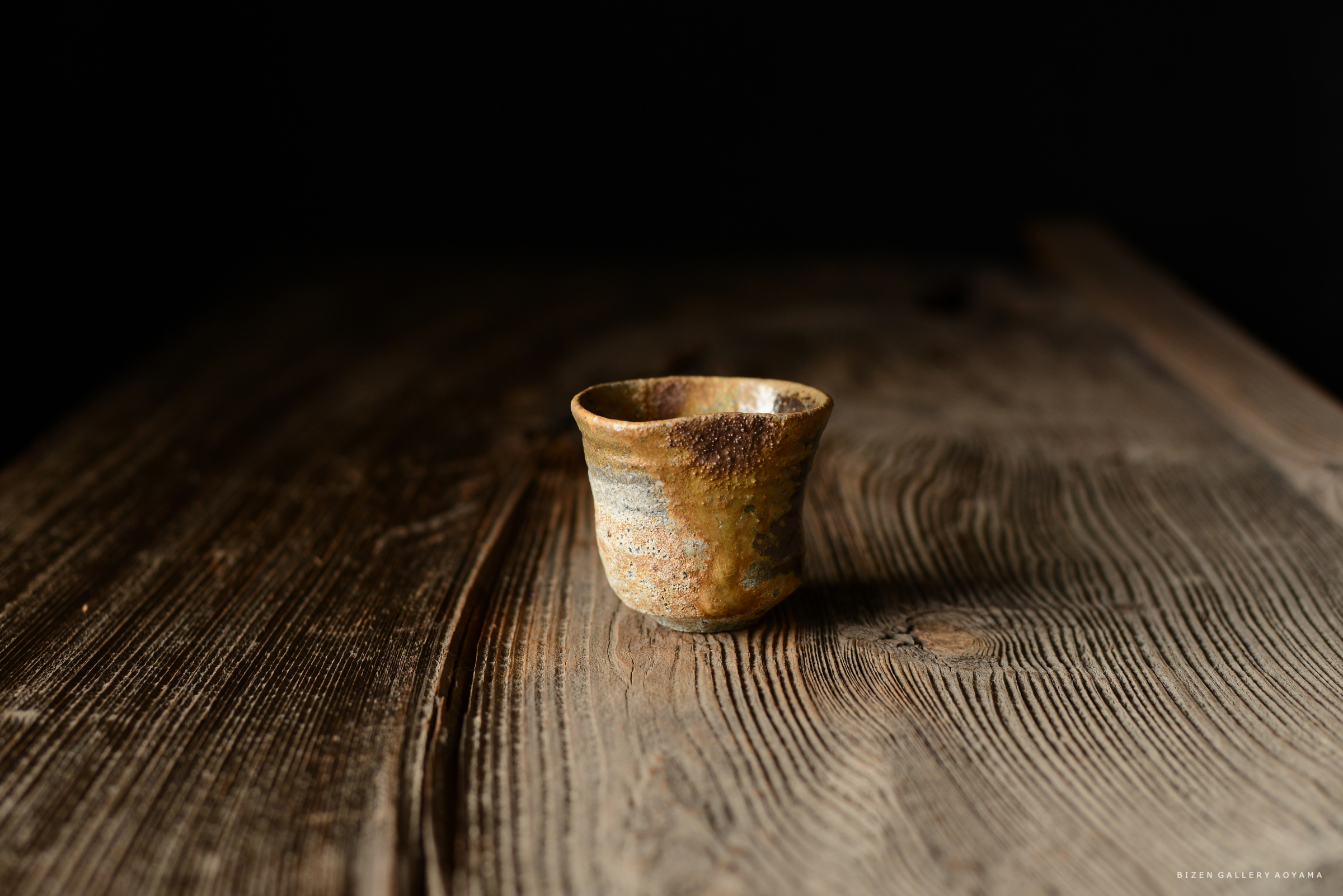 黒い背景の前に置かれた、陶器の小さなカップ。カップは複雑な色合いと質感を持ち、木のテーブルの上に置かれている。