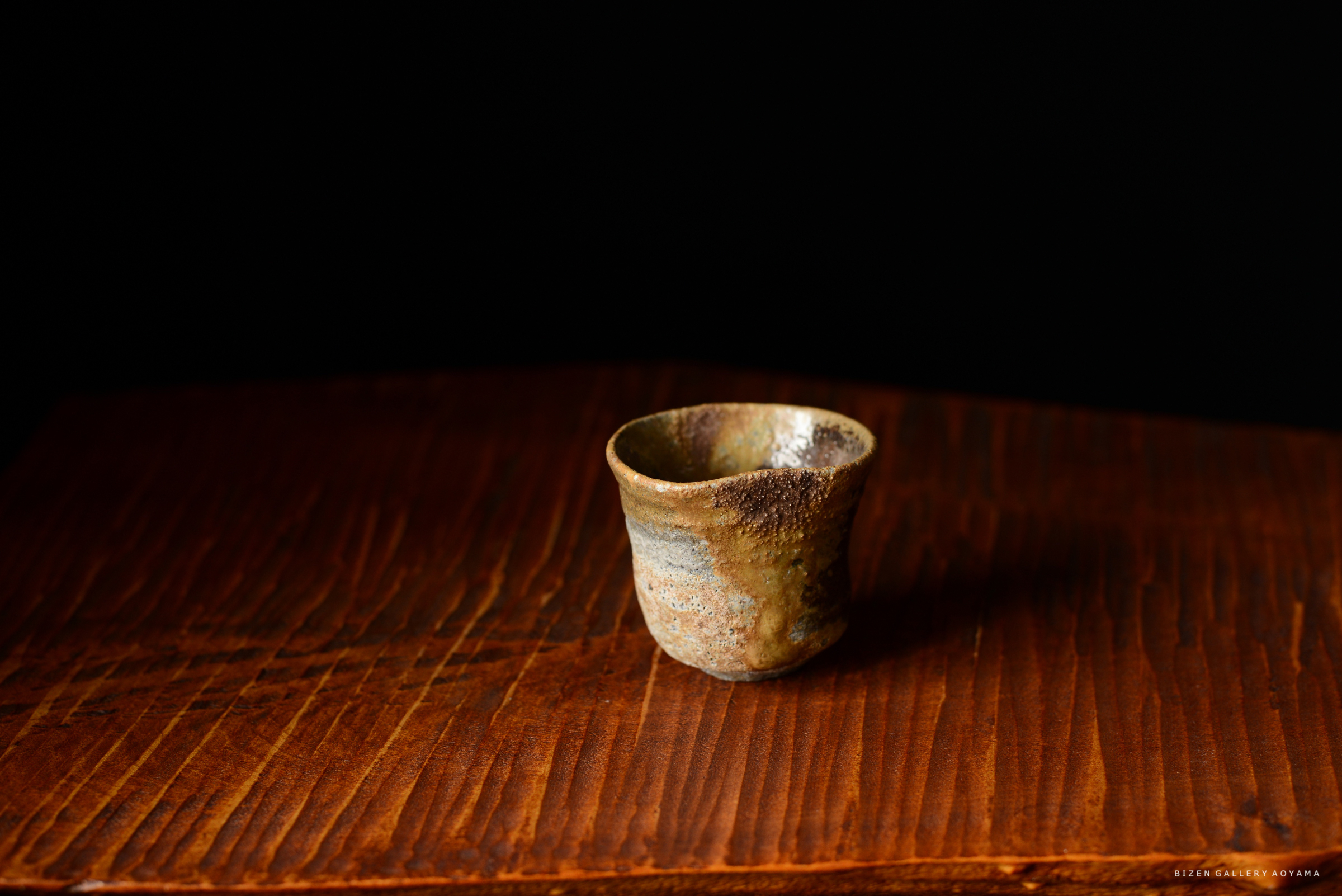 黒背景の上に置かれた焼き物の茶碗の画像。木製のテーブルの上にあり、独特な形状と色合いを持っている。