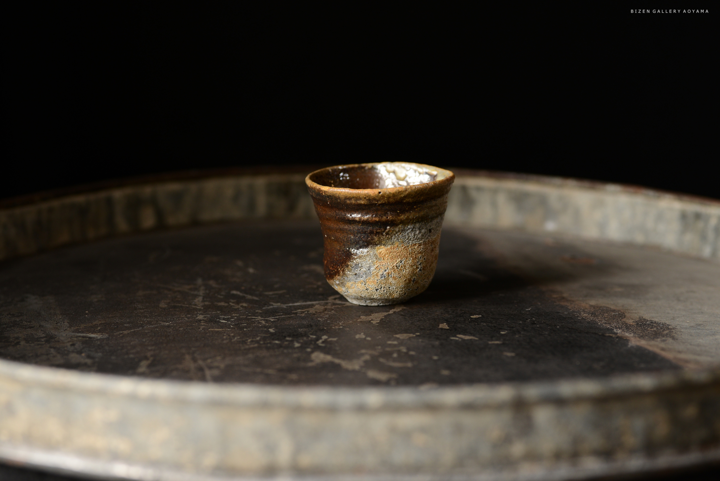 黒い背景の上に置かれた小さな土器のような壺。銀色のトレーの上にあり、質感や色合いが強調されている。