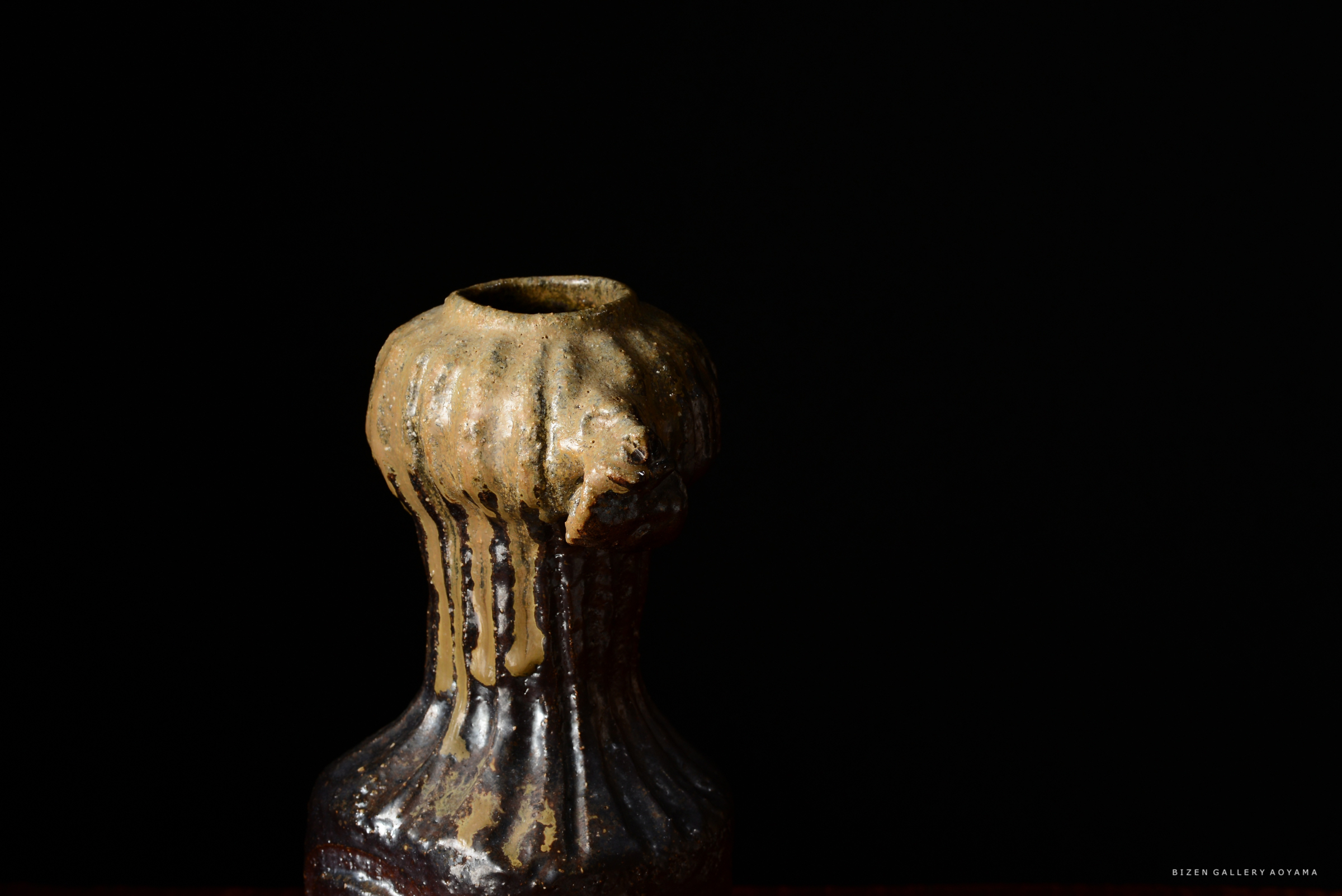 黒い背景の前に置かれた、独特な形状の陶器の壺。上部から滴る釉薬が特徴です。