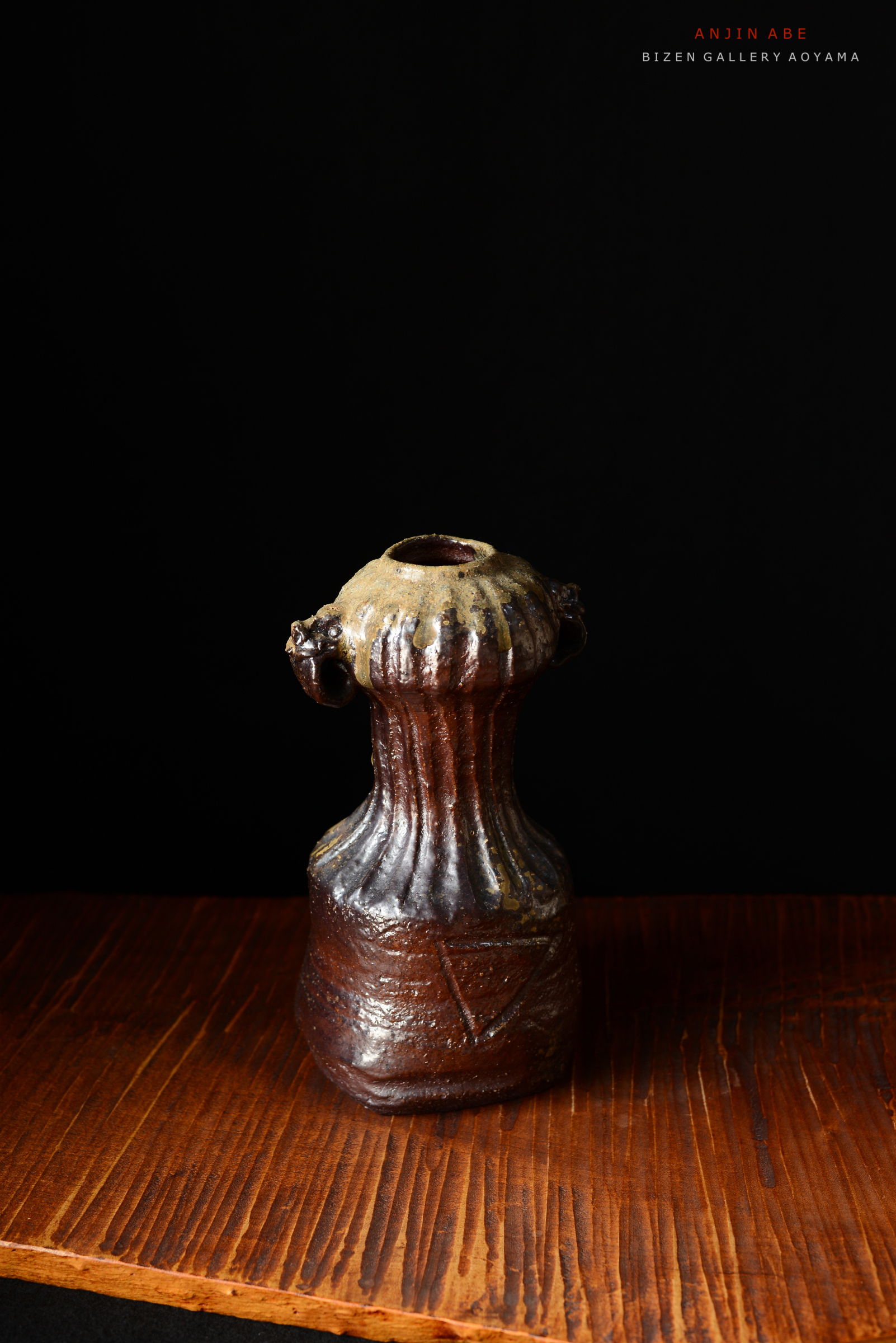 黒い背景の前に置かれた陶器の壺。細長い首と波状の装飾が特徴で、取っ手部分には顔のようなデザインが施されている。木製の板の上に置かれている。