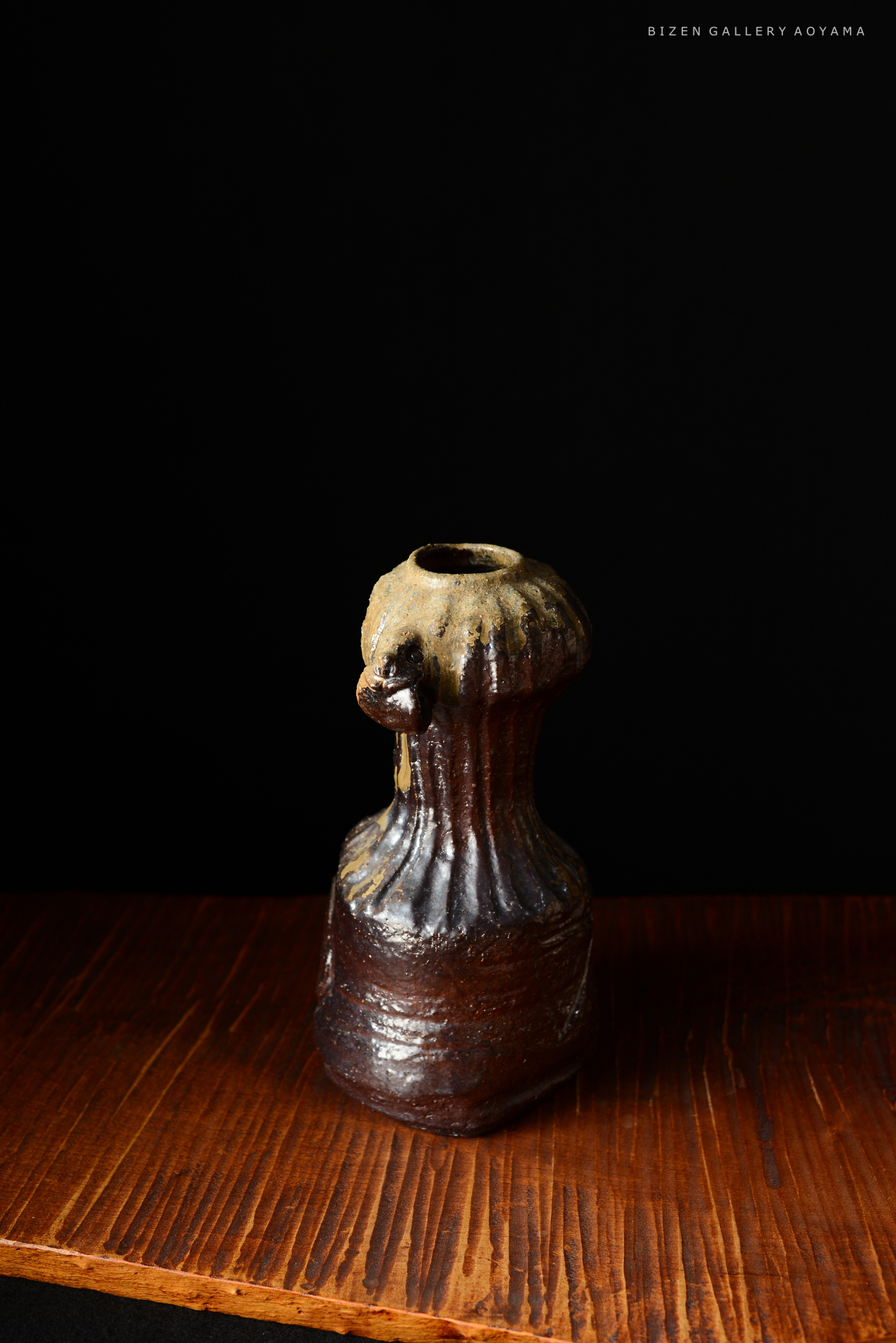 黒い背景に置かれた、独特な形状の備前焼の陶器。木製の台の上にあり、上部はやや丸く、下部は細長いデザインを持つ。