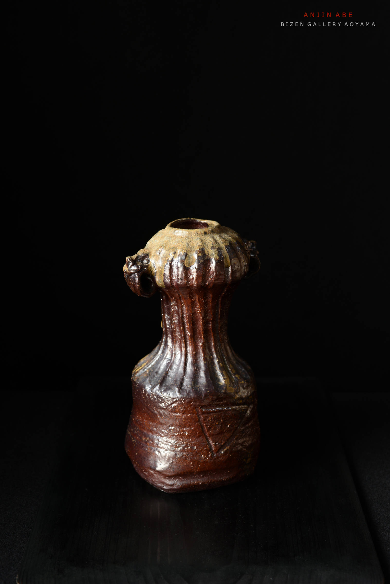 黒い背景に置かれた、細長い形状の焼き物。上部に装飾が施されており、口が広がっている。