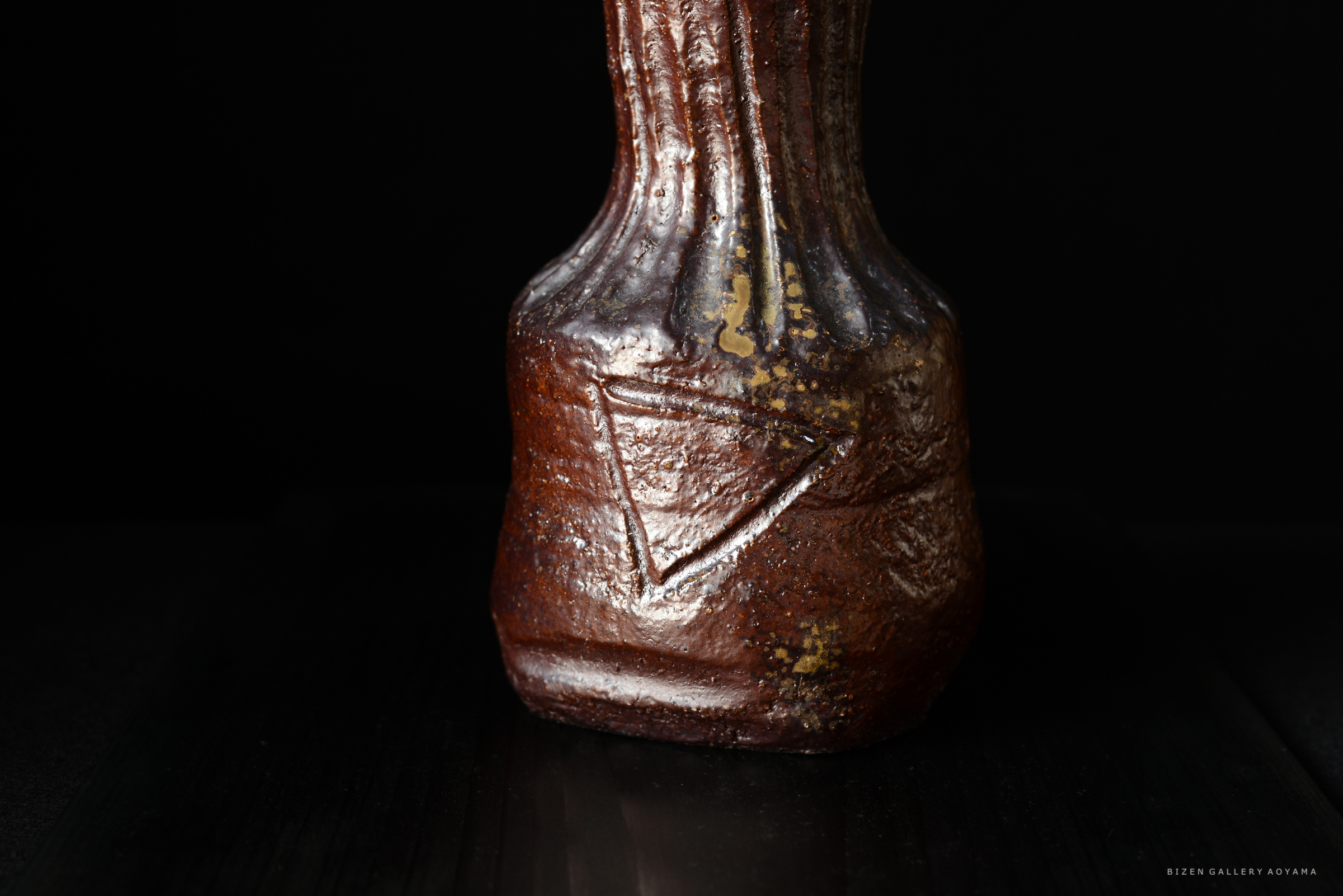 黒い背景に置かれた茶色の陶器の壺。表面には三角形の模様が刻まれている。