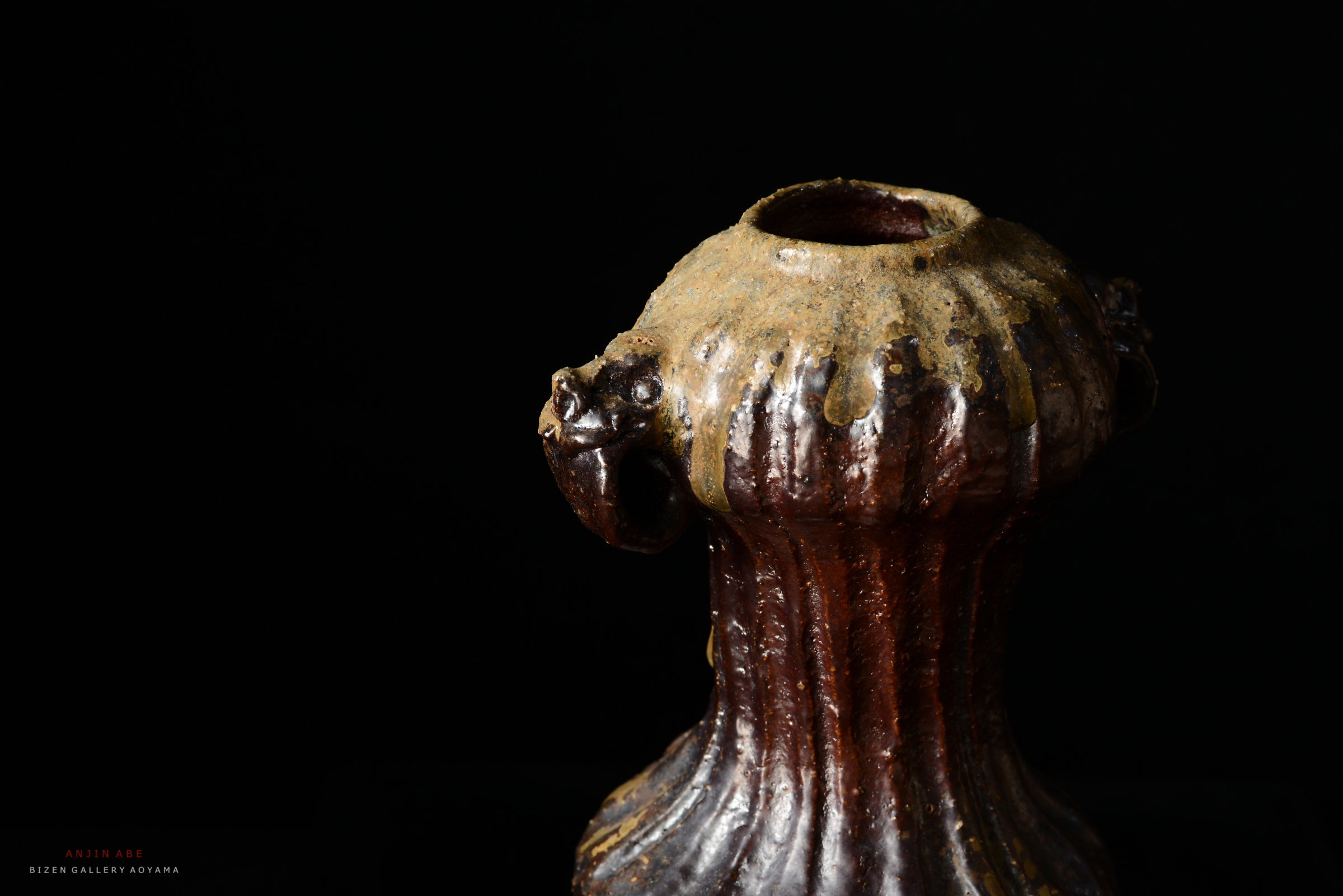 黒い背景の前に置かれた古代の陶器。上部に特徴的な装飾が施された形状を持ち、色合いは茶色と金色が混ざり合った印象。