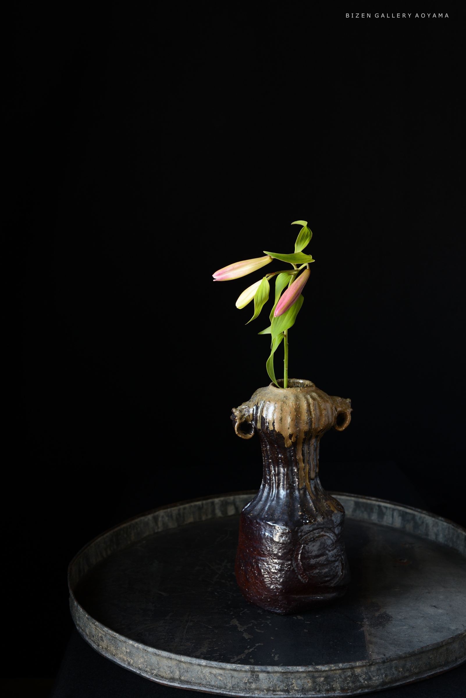 黒い背景の上に置かれた茶色の陶器の花瓶に、ピンクのつぼみを持つ花が生けられているシンプルなアレンジメント.