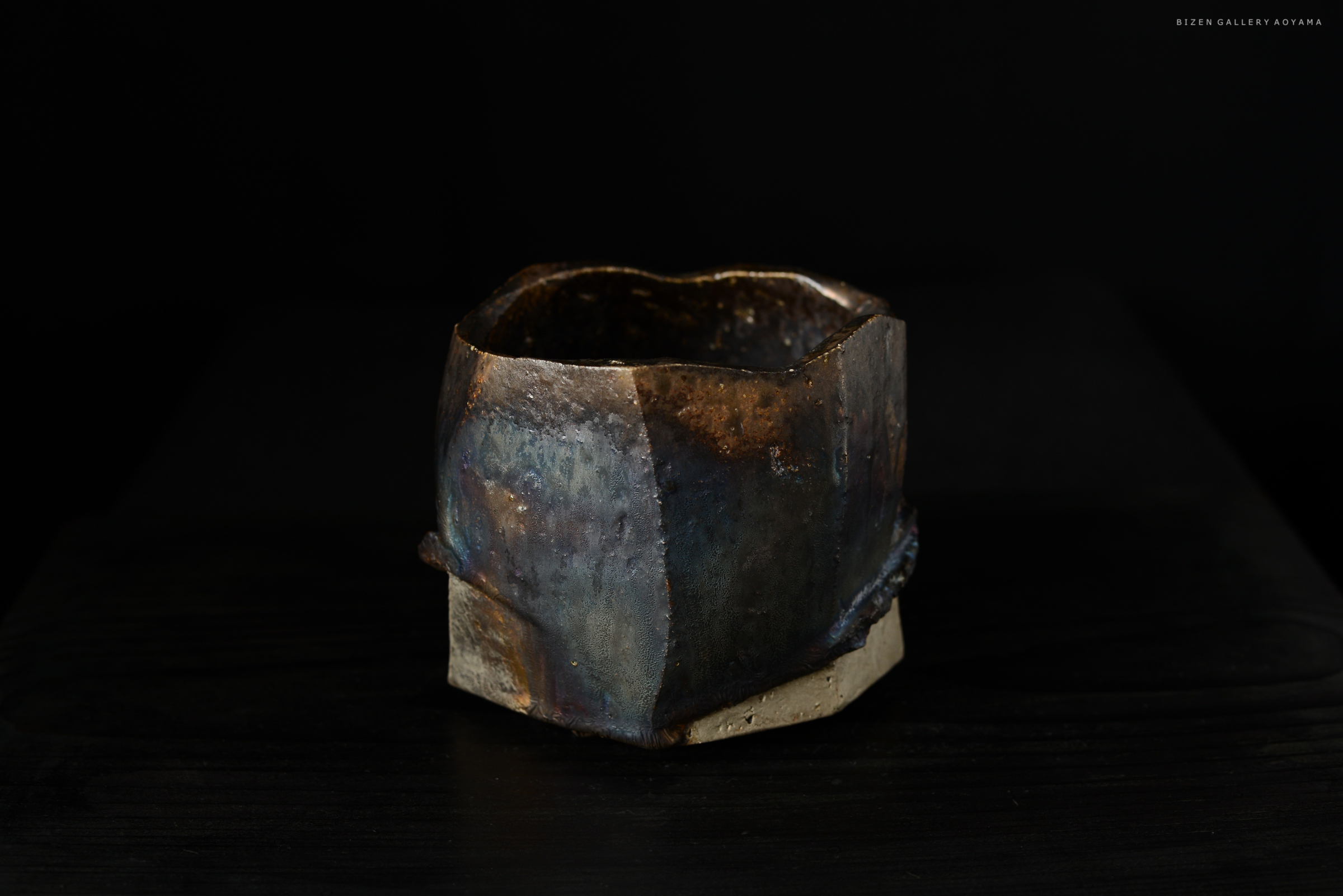 黒い背景の上に置かれた独特な形の陶器の容器。表面には青や茶色の色合いが見られ、焼き物の技術による独自の模様がある。