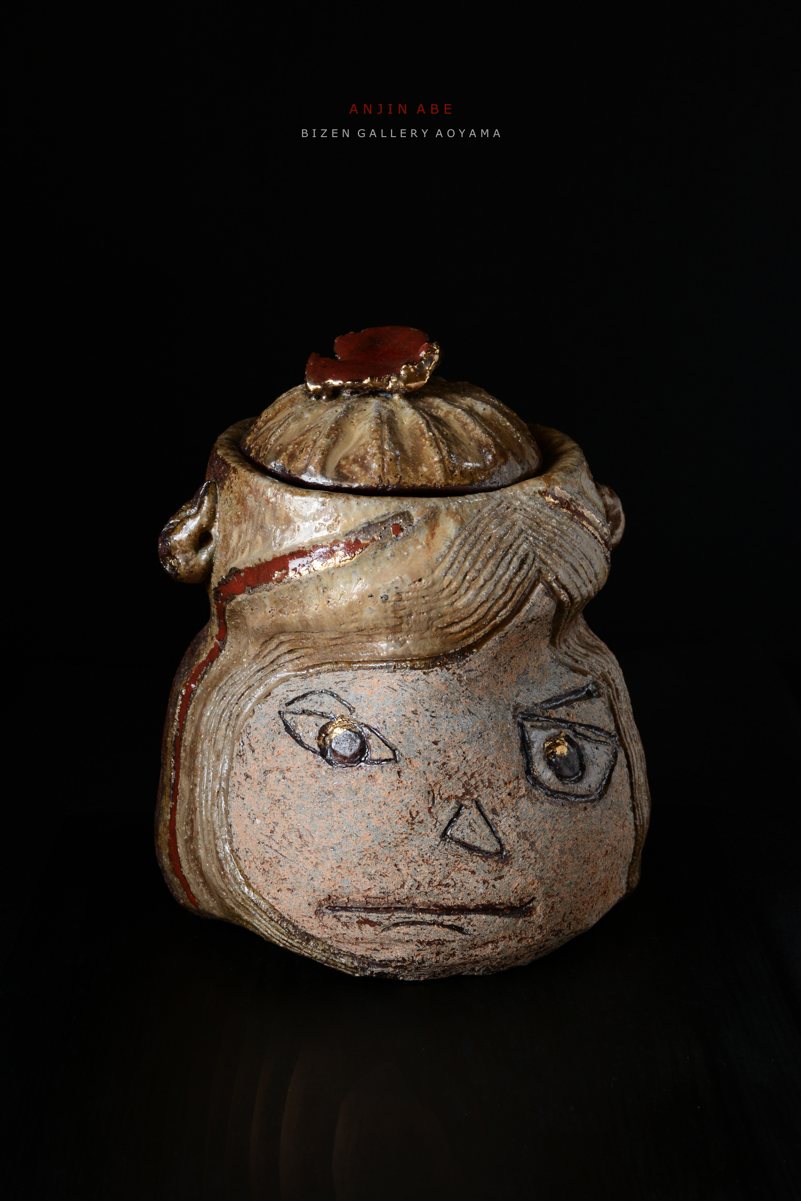 アート作品の陶器の容器。顔の表情が描かれたデザインで、蓋には赤い装飾が施されている。背景は黒。