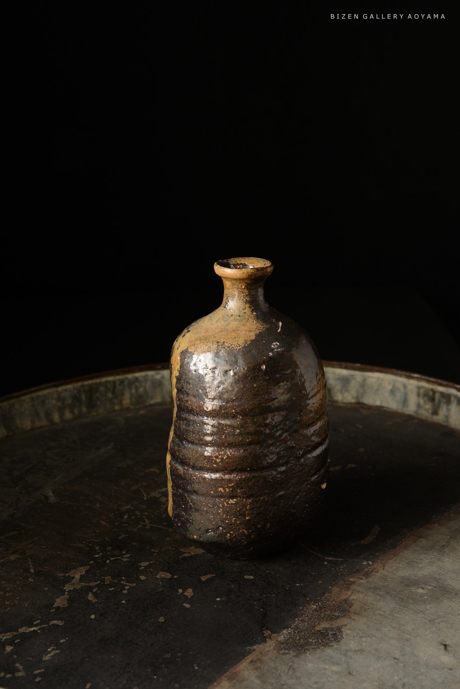 黒い背景の前に置かれた、ビゼン焼きの陶器の瓶。表面は茶色と黒の釉薬で覆われ、独特の質感があります。