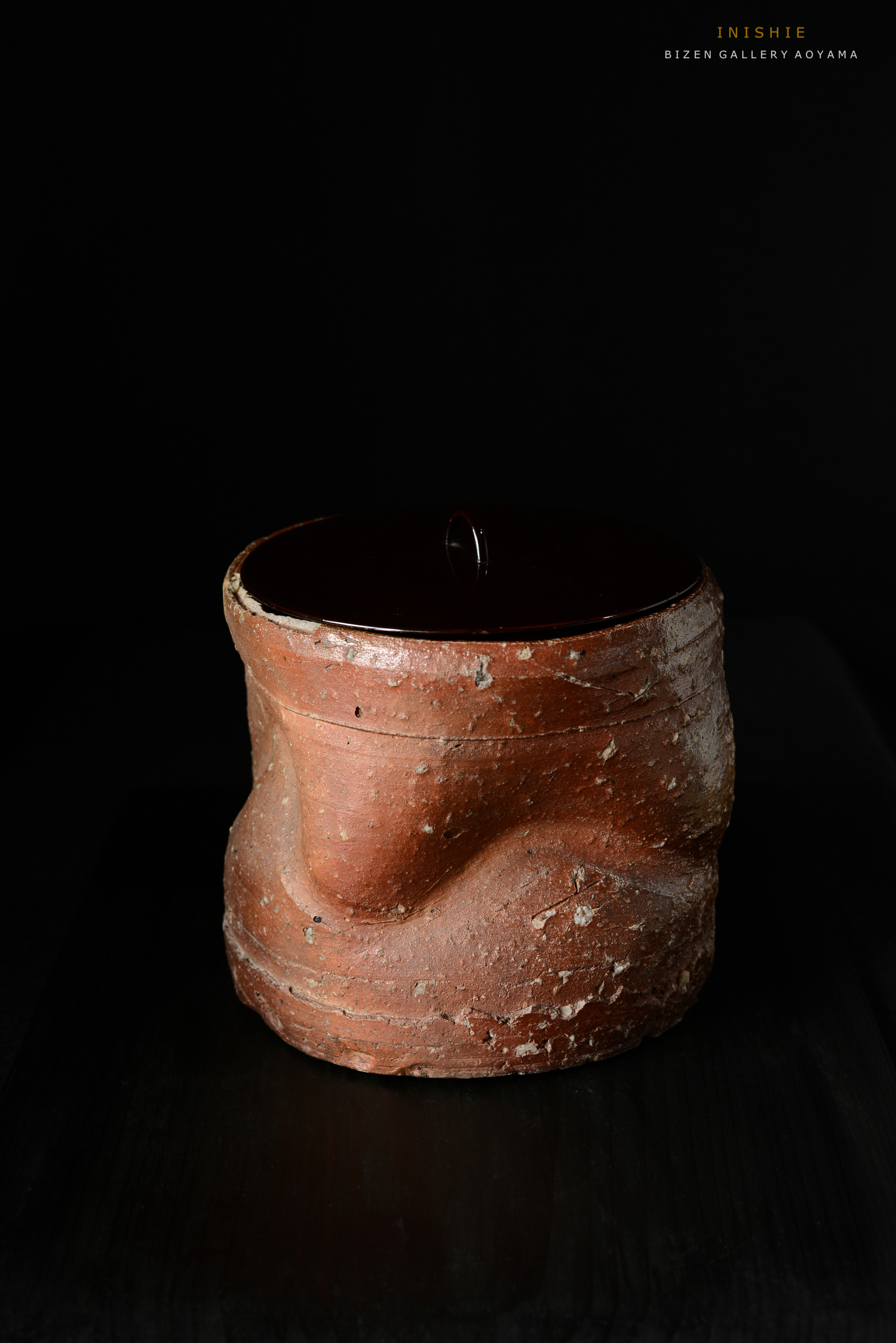 黒い背景の上にある陶器の壺。表面はざらざらしており、上部には光沢のある蓋が載せられている。