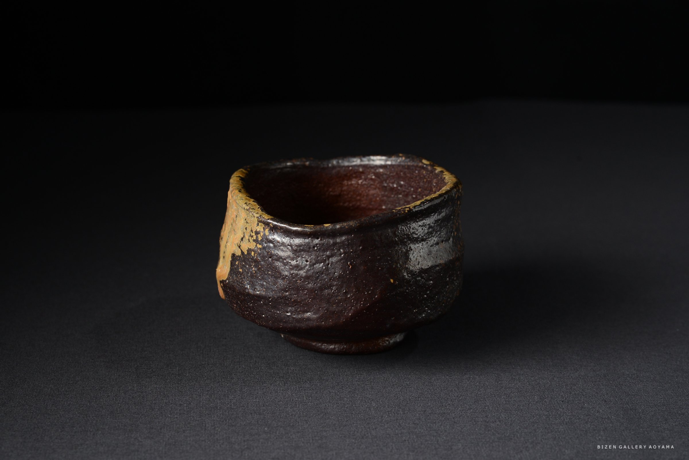 黒い背景の上に置かれた、独特な形状の茶碗。茶碗の側面には、土の色合いが見られ、口元は金色の縁取りが施されている。