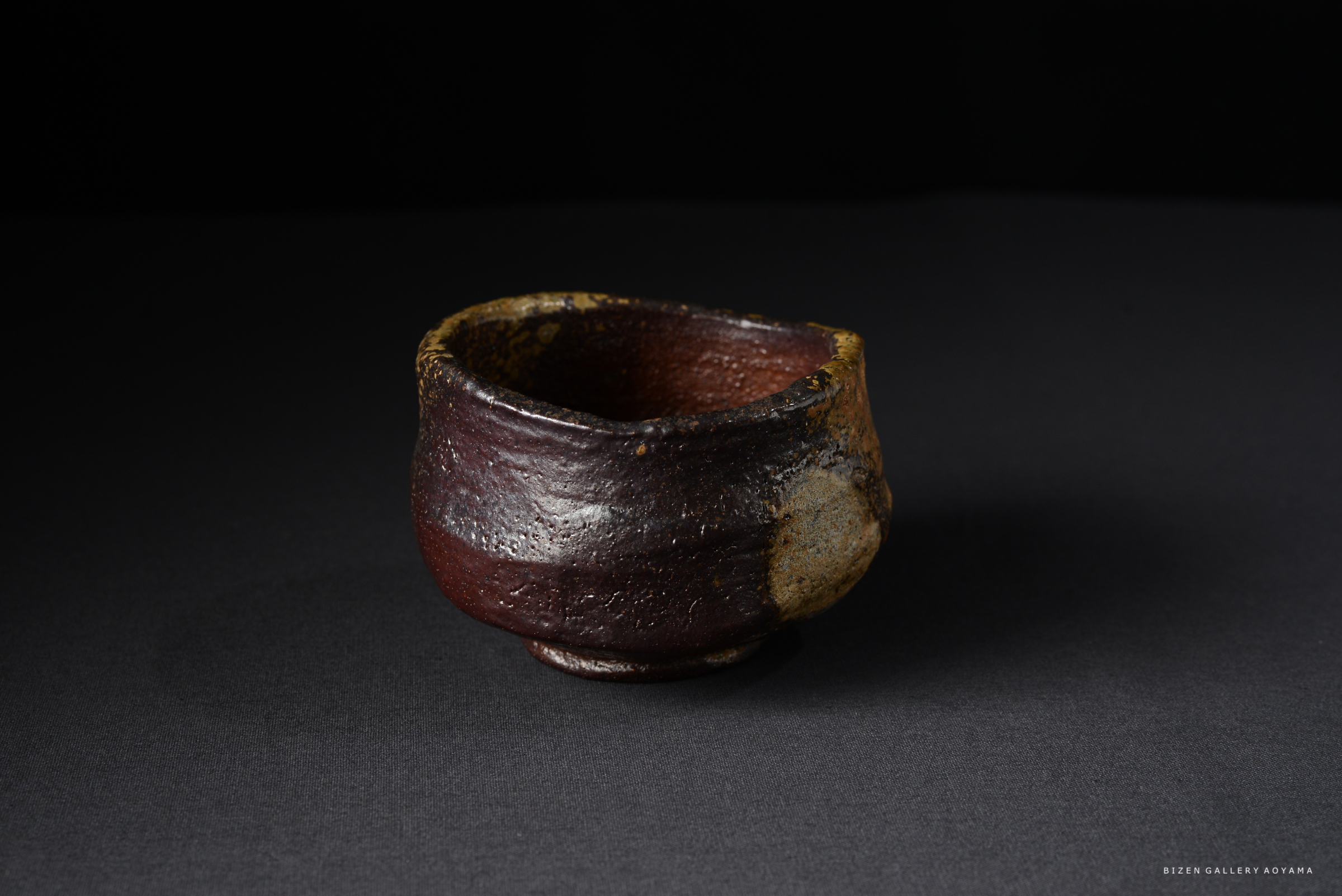 黒い背景に置かれた備前焼の茶碗。複雑な色合いと質感を持ち、力強い造形が特徴的。