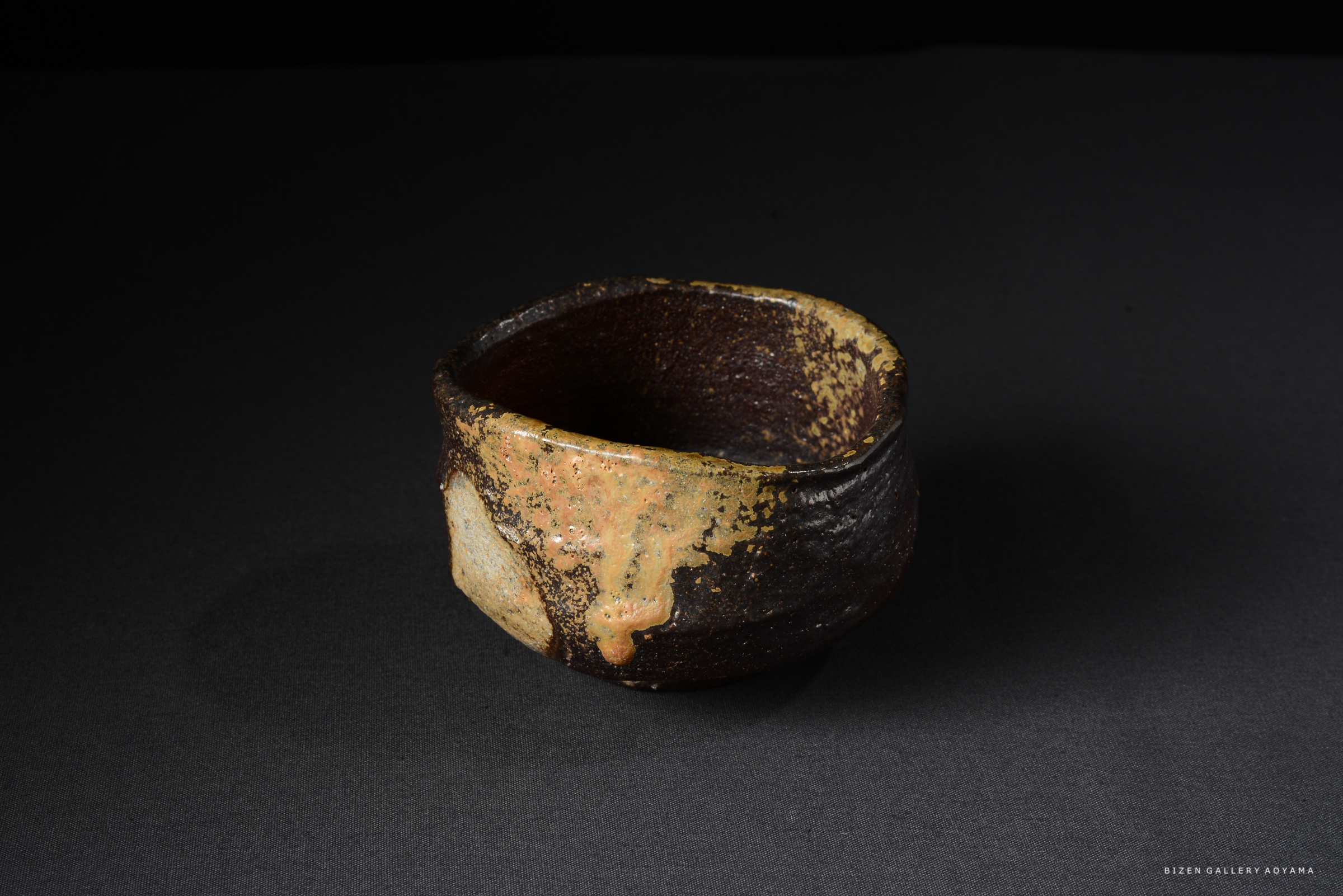 黒い背景の上に置かれた、独特な形状の陶器の茶碗。外側は茶色で、所々に金色と黒色の釉薬がかかっている。
