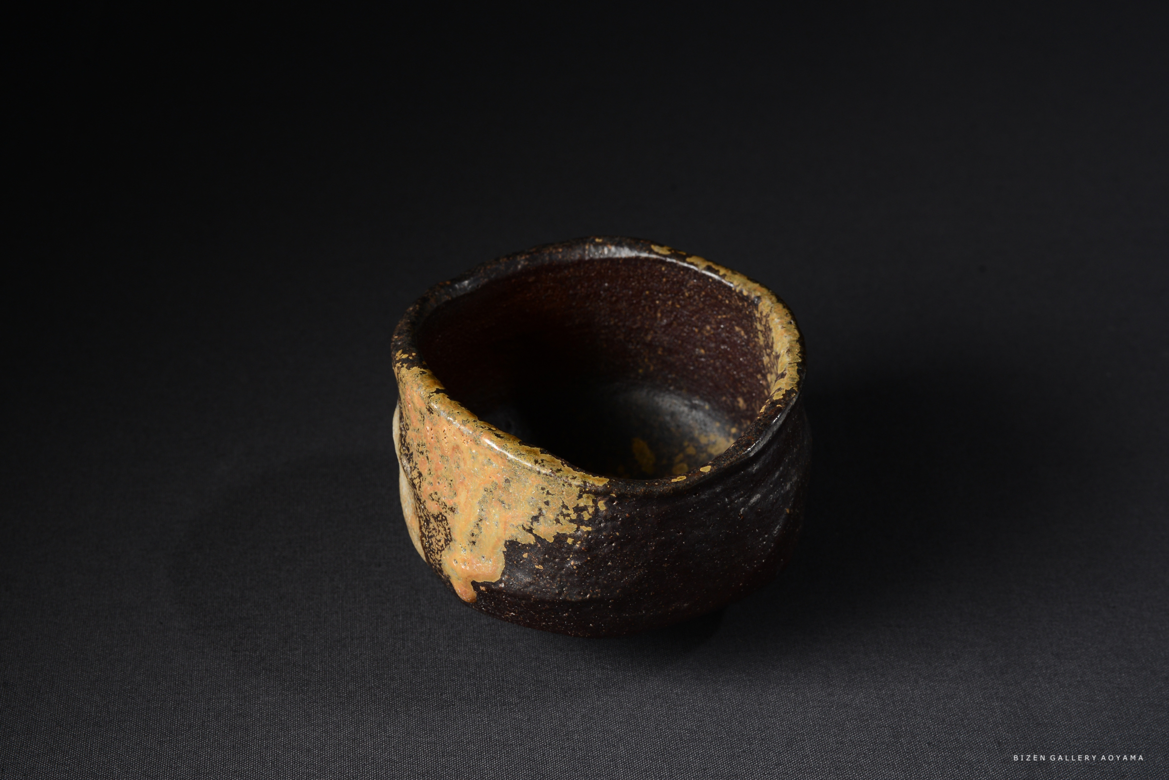黒い背景に置かれた独特な形状の陶器の器。外側は茶色と黒の不均一な色合いで、内側は深い黒色。