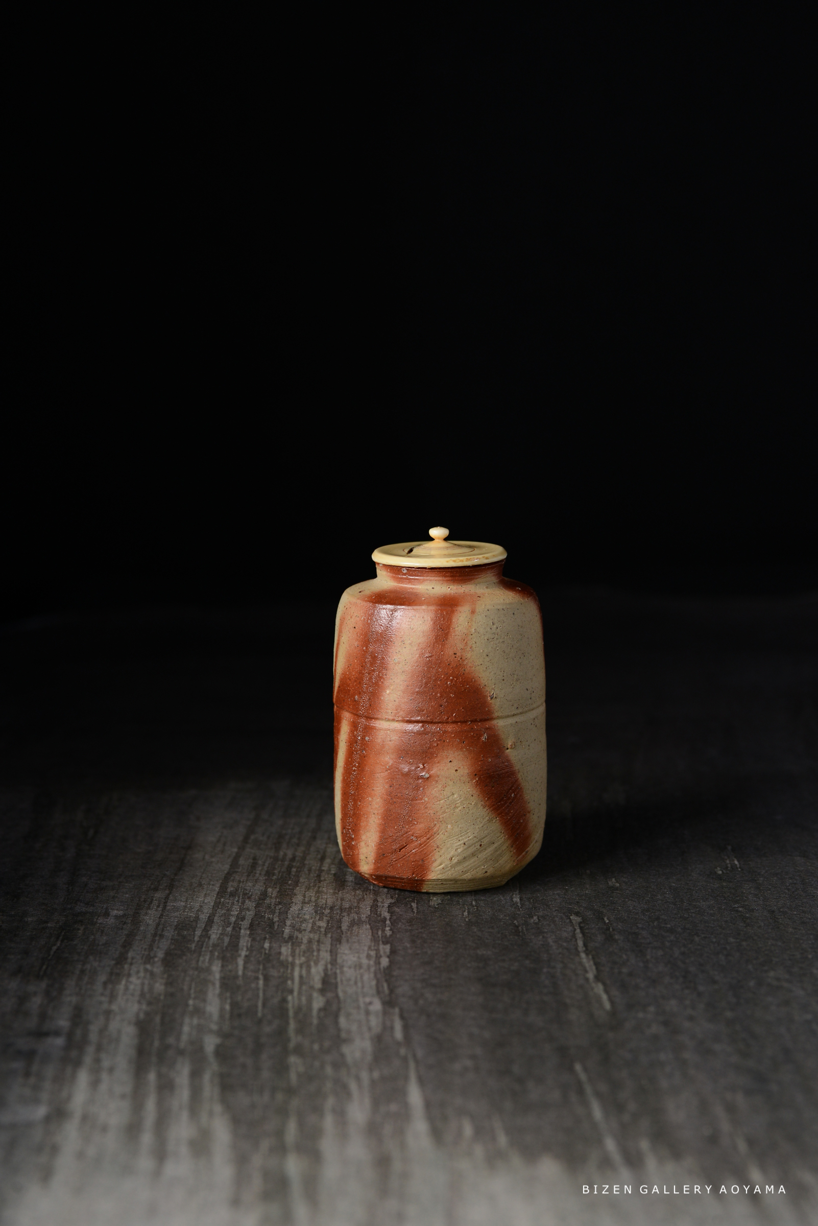 木の蓋が付いた、美しいビゼン焼の陶器の壺。暗い背景の上に置かれた、赤い模様の入った自然な色合いが特徴。