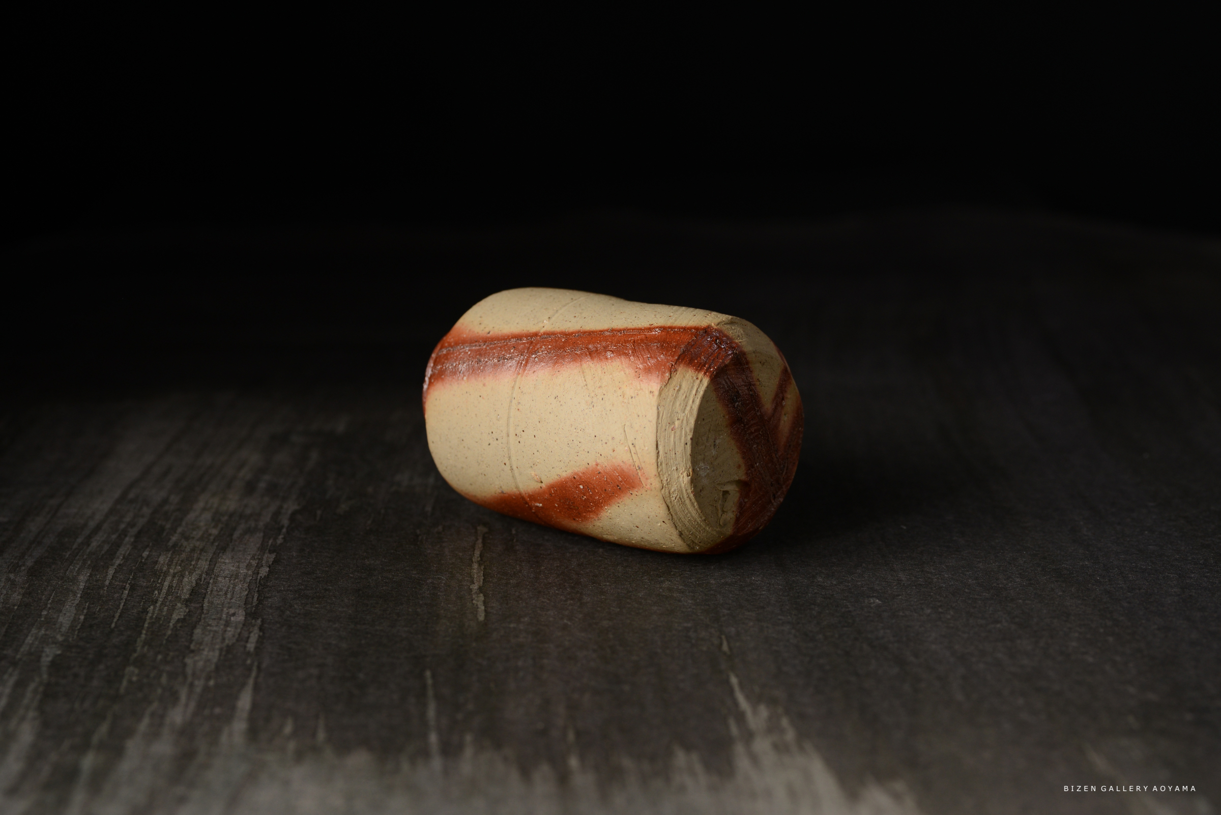 素焼きの陶器で、赤い模様のある円筒形のオブジェ。黒い背景の上に置かれている。