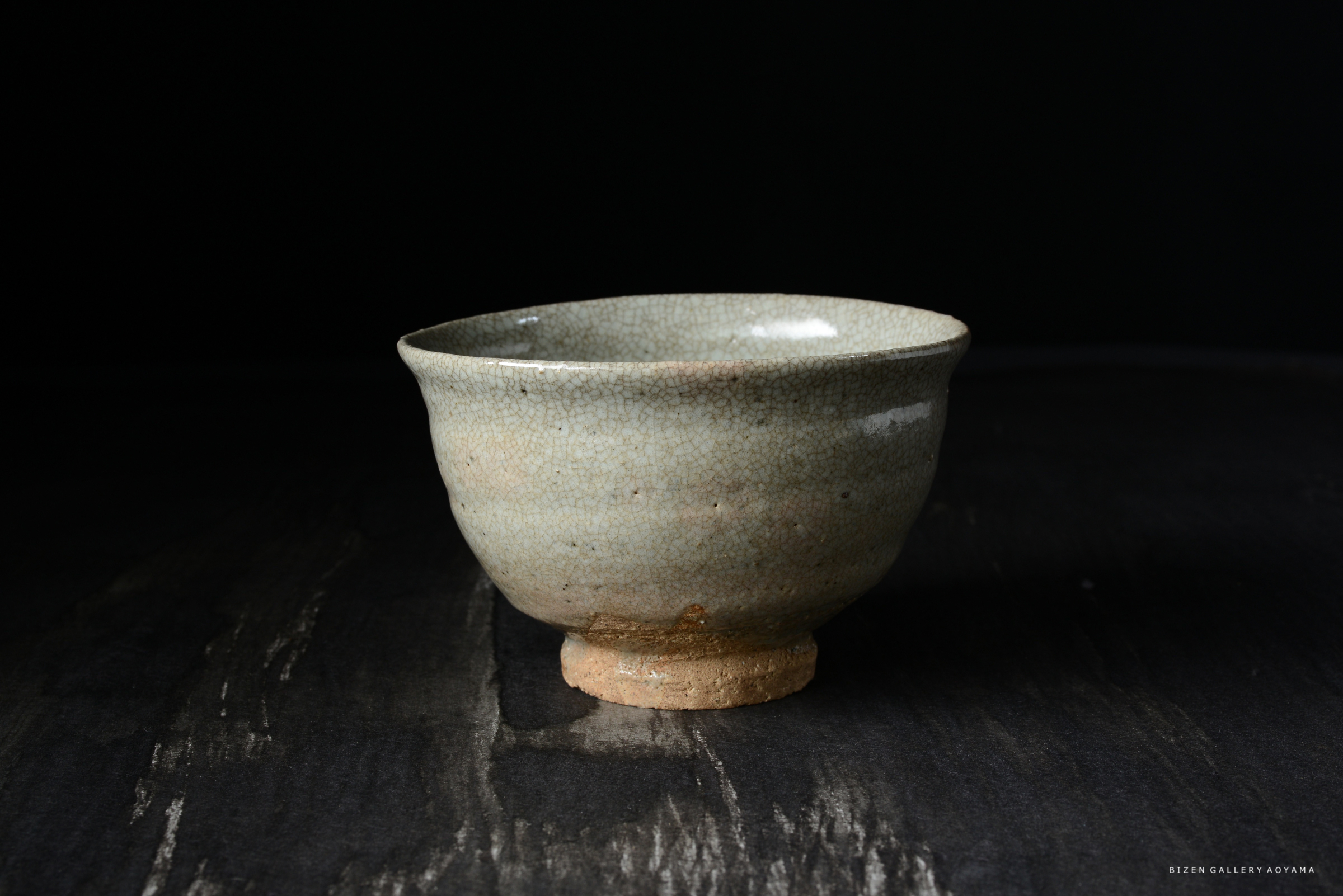 薄い青緑色の釉薬が施された陶器の茶碗。底は素焼きの土でできており、うっすらとしたひび模様が見える。暗い背景に置かれたシンプルなデザイン。